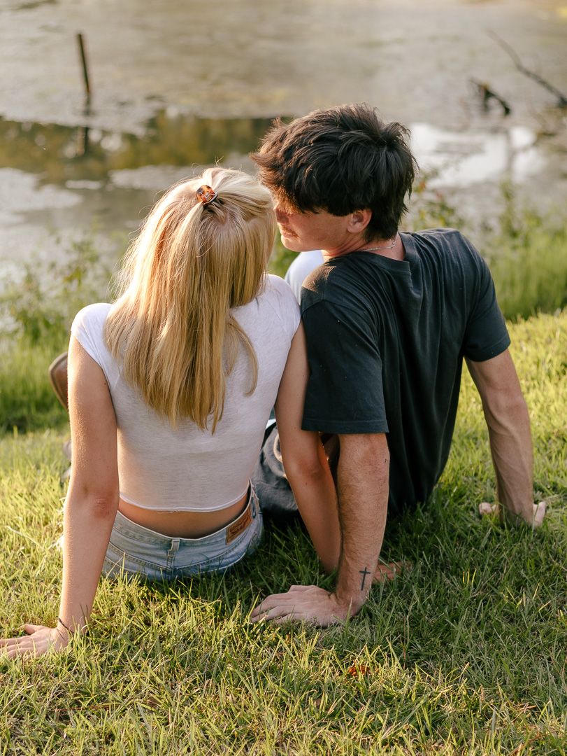 Young couple sits close together on a grassy lakeside bank, facing the water and sharing a quiet, romantic moment in warm sunlight.