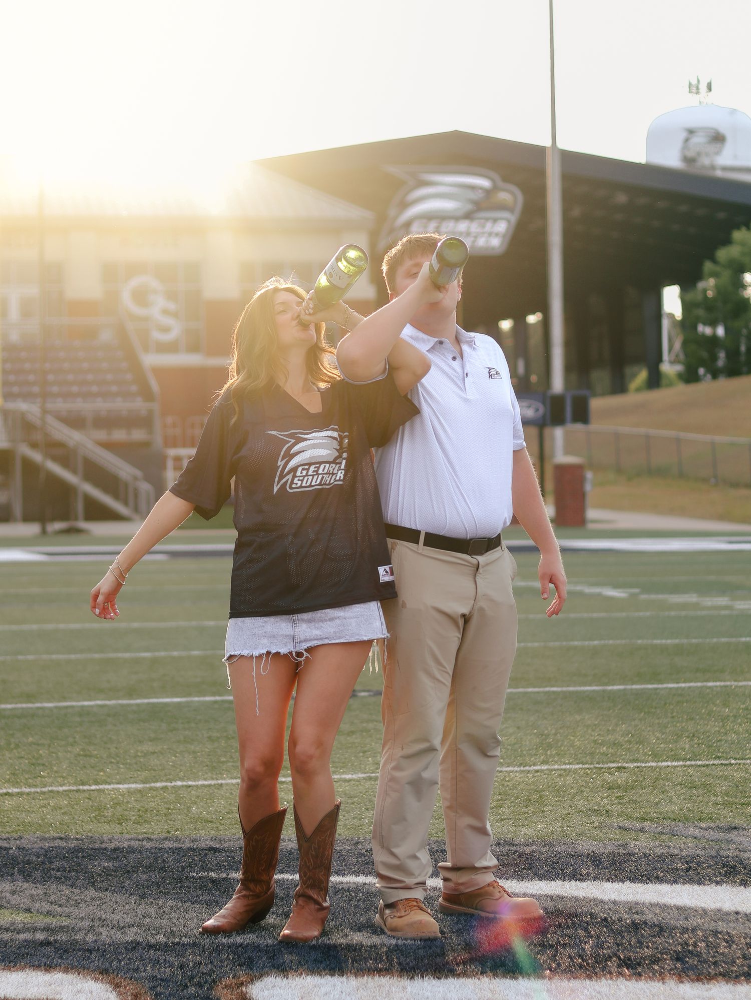 Couple in Georgia Southern gear toast champagne bottles on a football field at sunset, with stadium seating in the background.