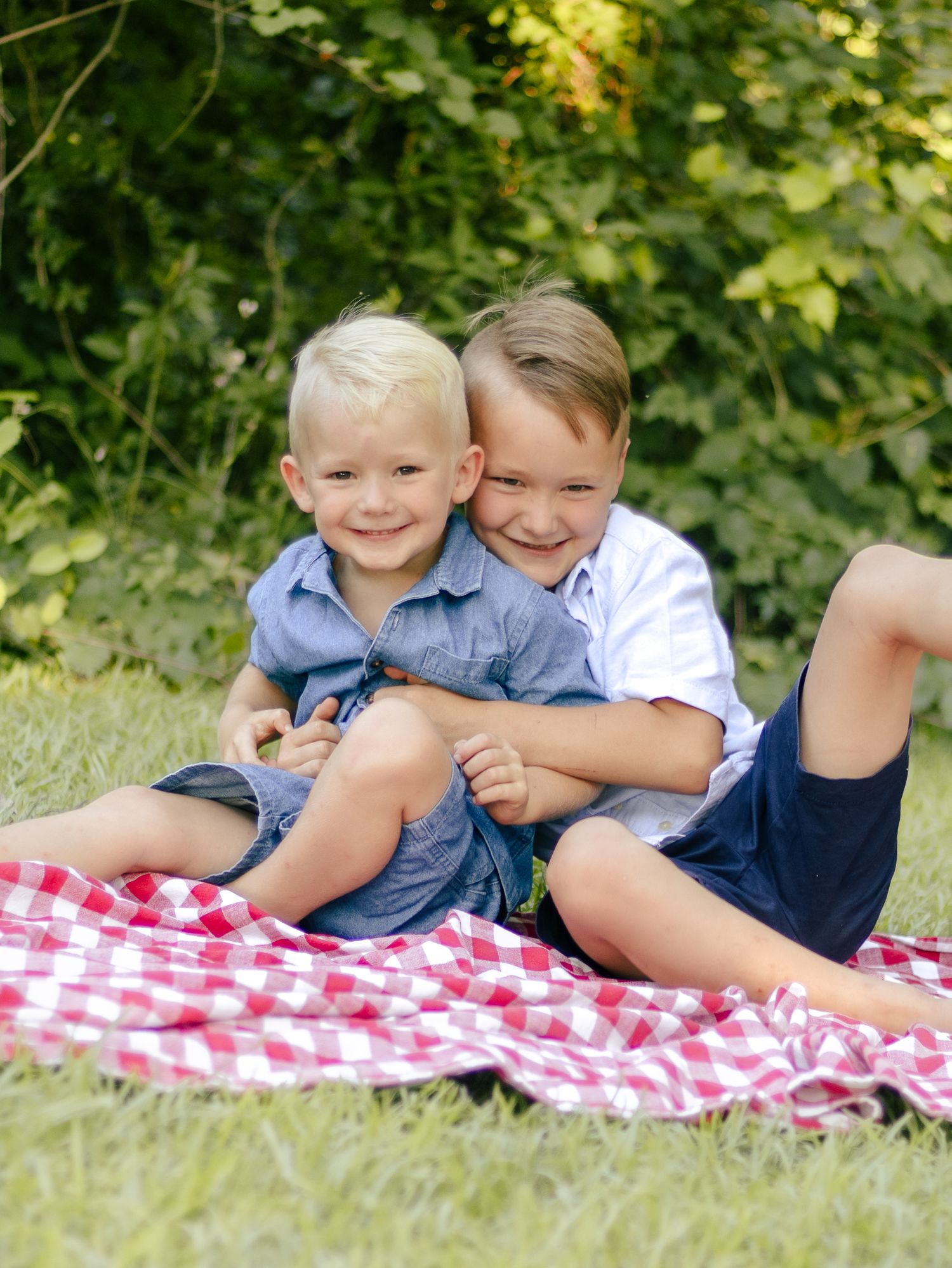 Two smiling boys in casual summer clothes hug and sit together on a red and white checkered picnic blanket on green grass.