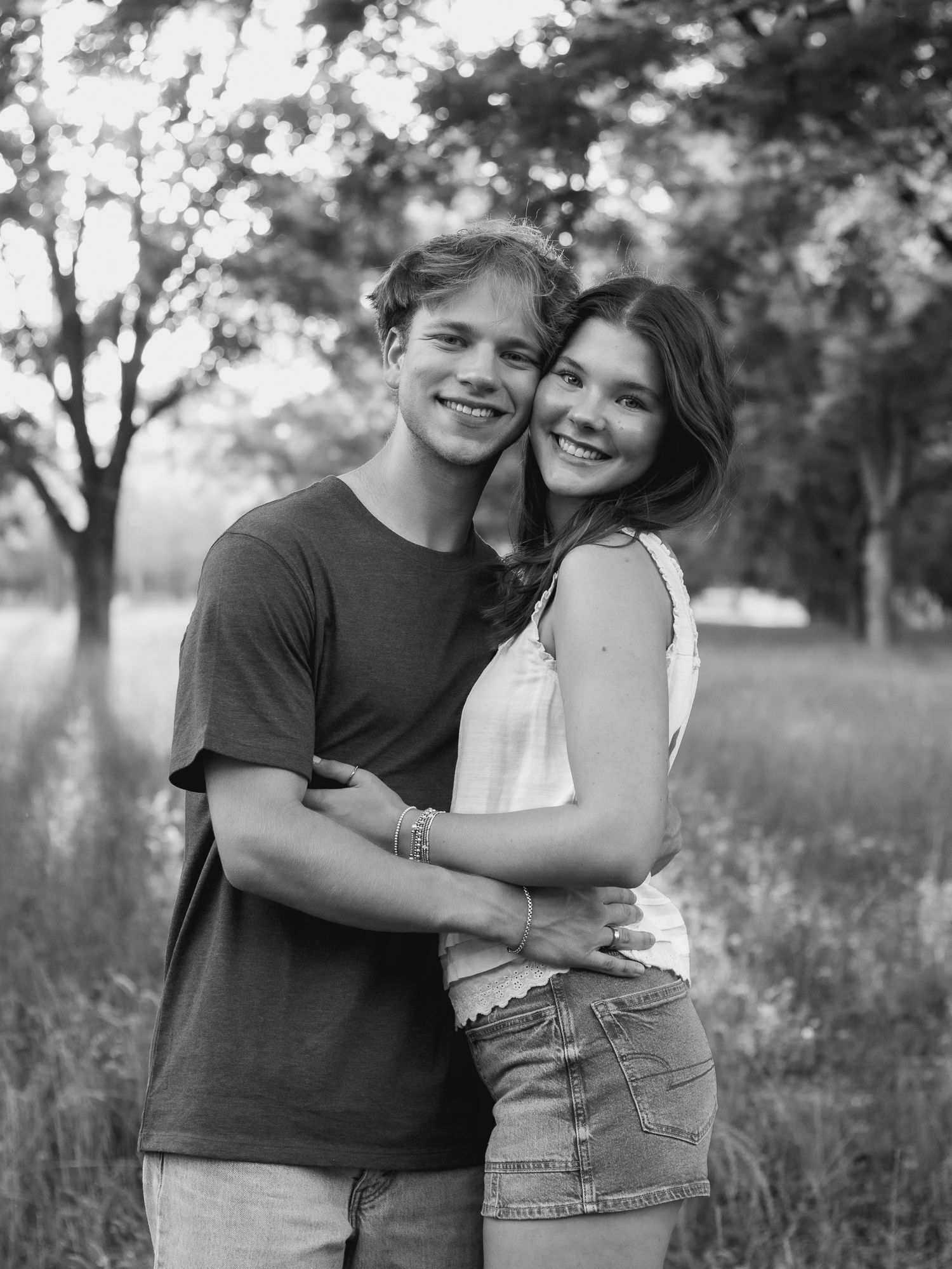 Smiling young couple embracing in a grassy field surrounded by trees in a black and white outdoor portrait.