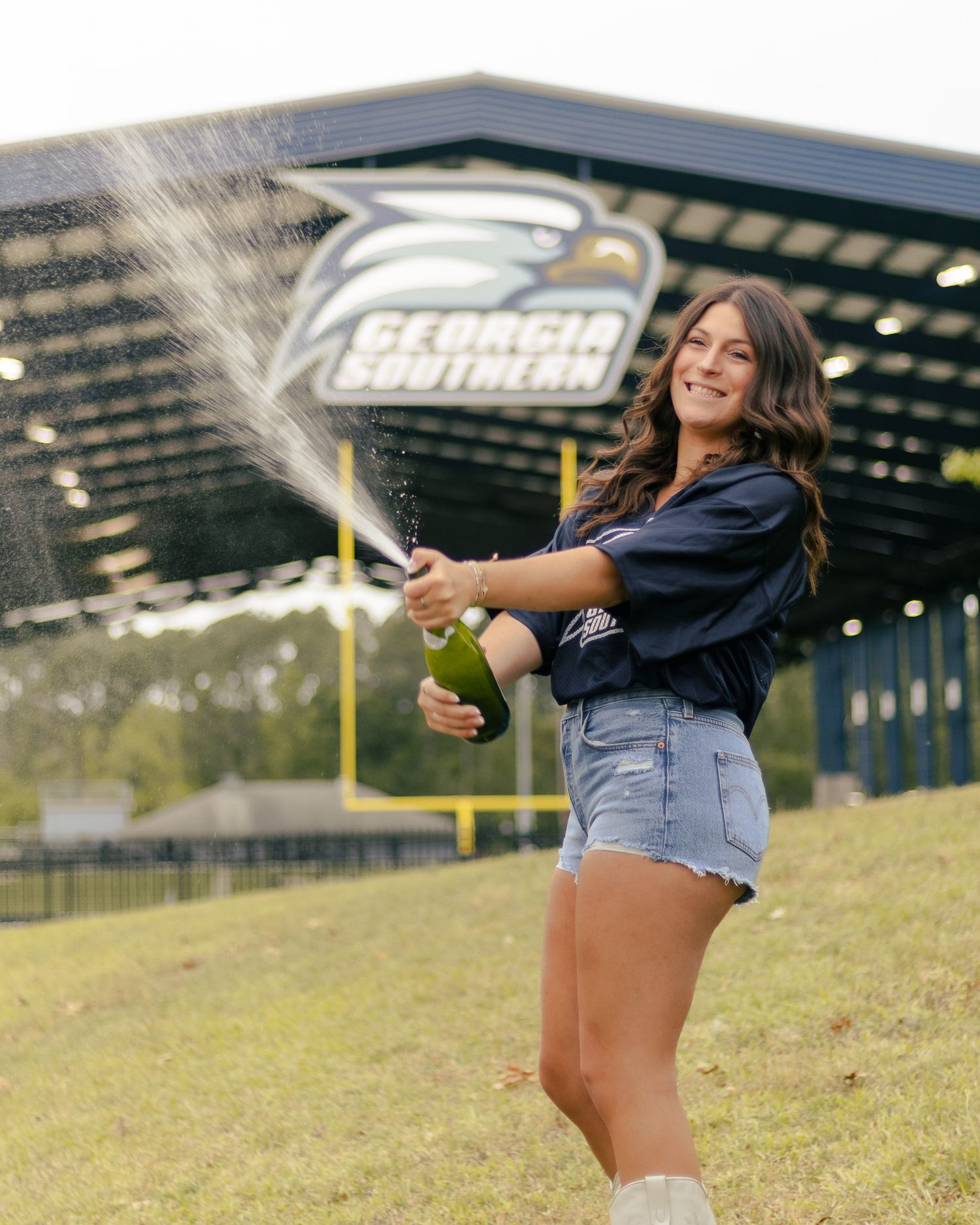 Smiling woman in denim shorts spraying champagne on a grassy hill outside the Georgia Southern stadium, celebrating graduation.