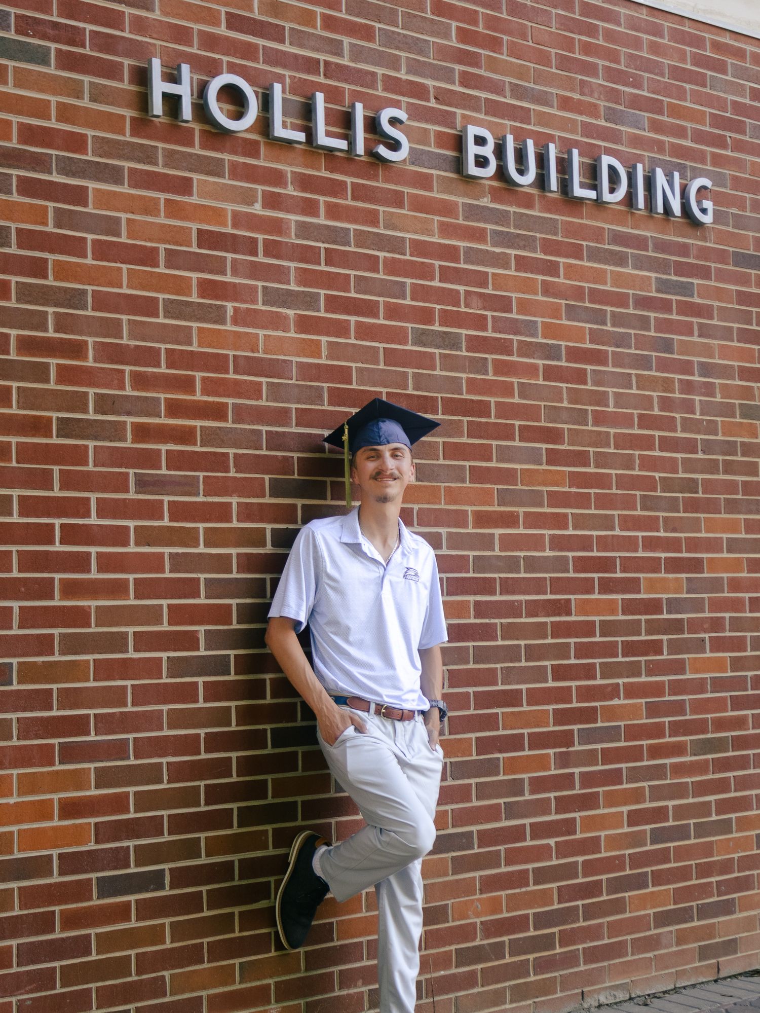 Graduate in a blue cap, white polo and khakis leaning on a red brick wall beneath the Hollis Building sign.