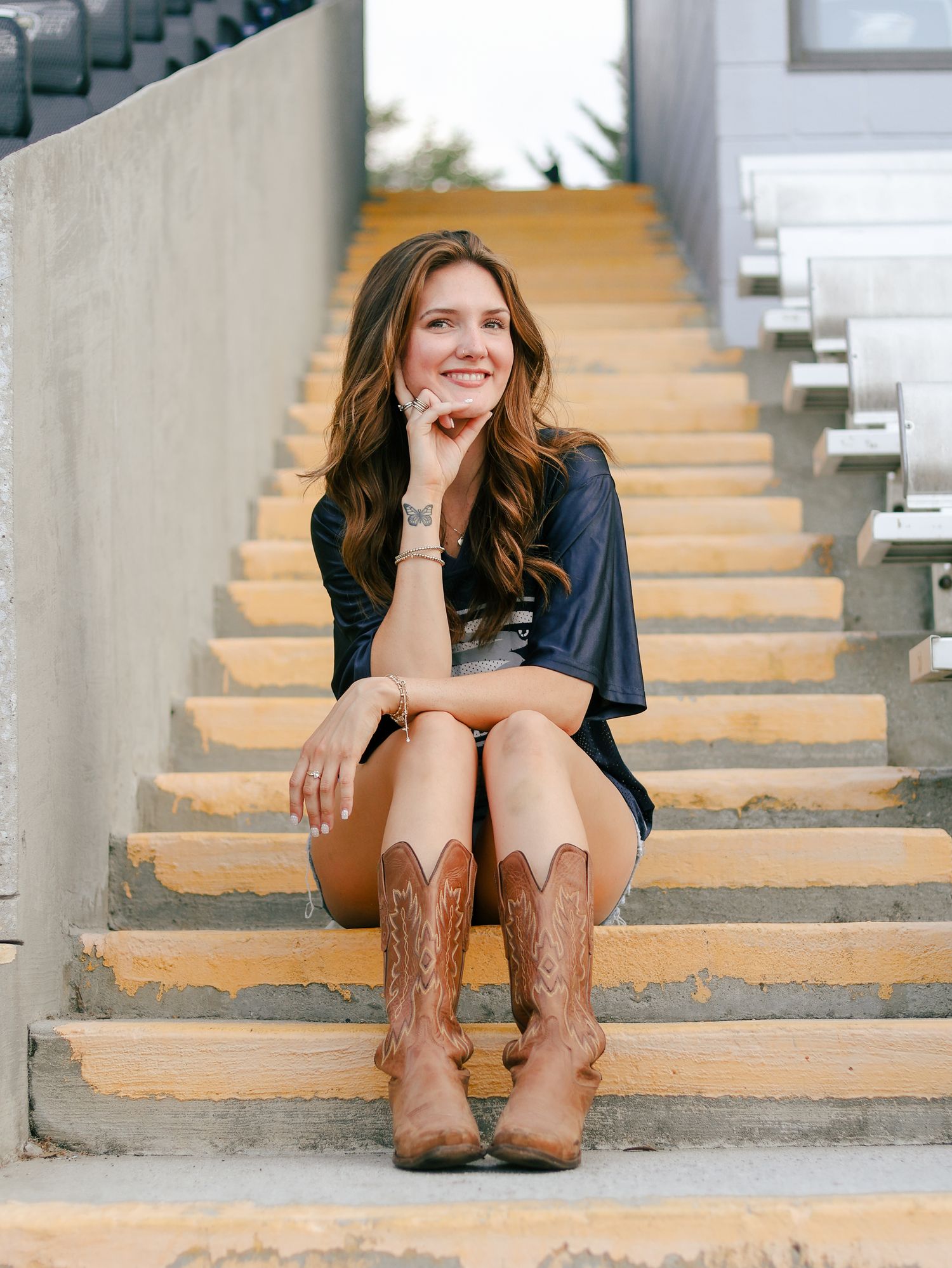 Smiling woman with long wavy hair sits on yellow-edged stadium steps in a navy top, denim shorts and tan cowboy boots.
