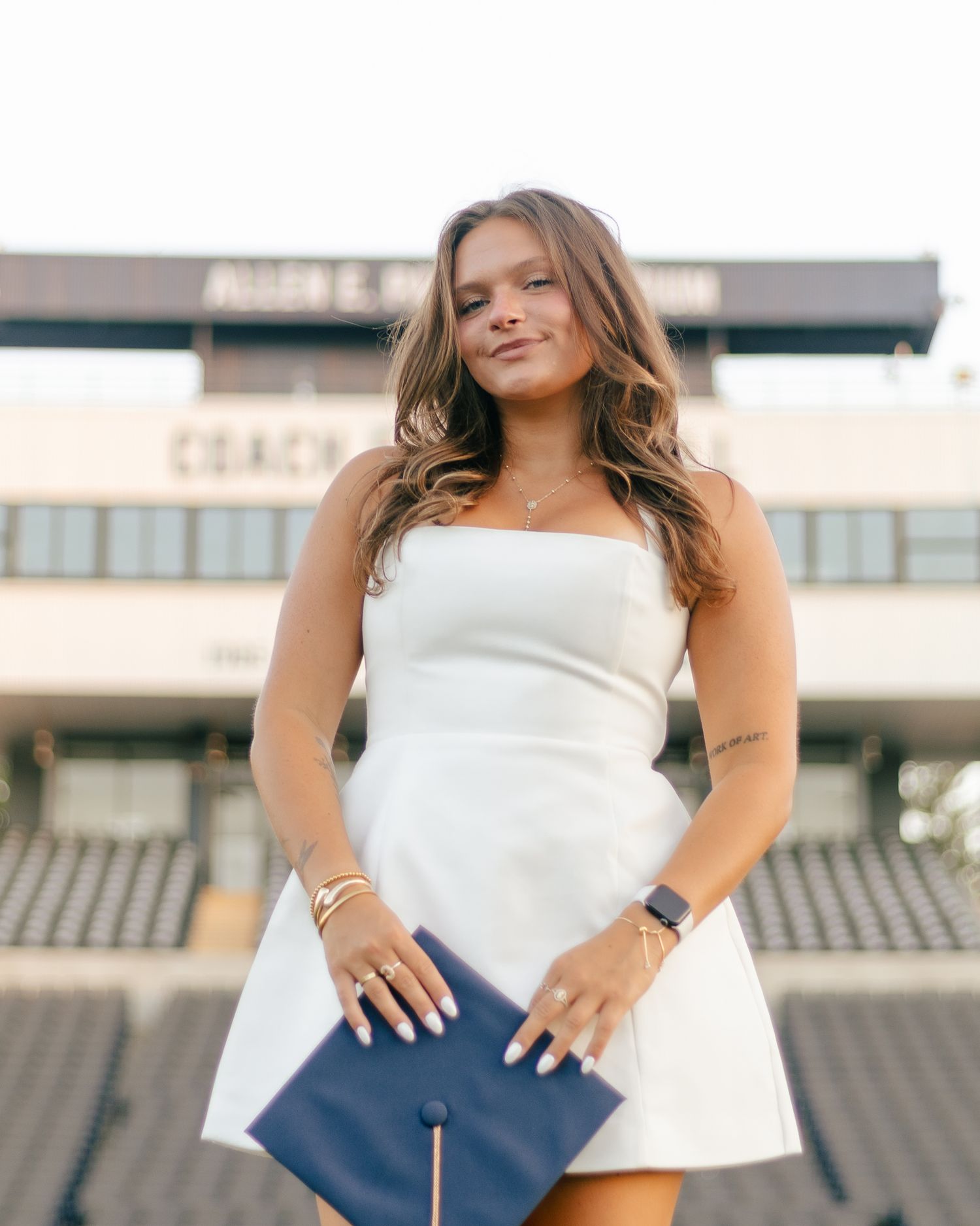 Smiling graduate in a white strapless dress holds a navy graduation cap while posing in front of a stadium grandstand at sunset.