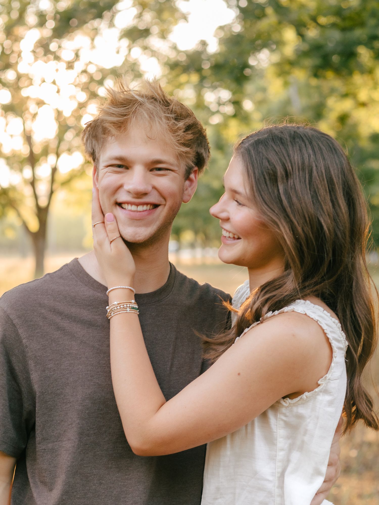 A smiling young couple embraces outdoors, the woman gently cupping the man's cheek, with sunlit trees softly blurred behind them.