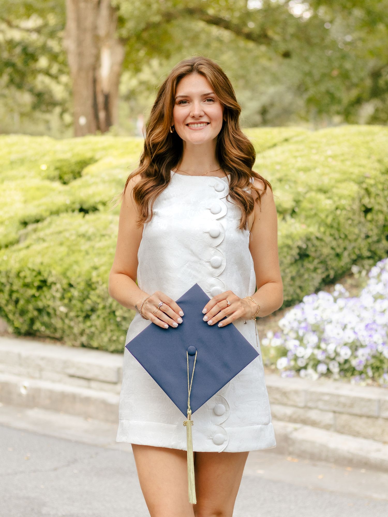 Smiling graduate in a white button-front mini dress holds a navy blue graduation cap with a tassel in front of green hedges and flowers.