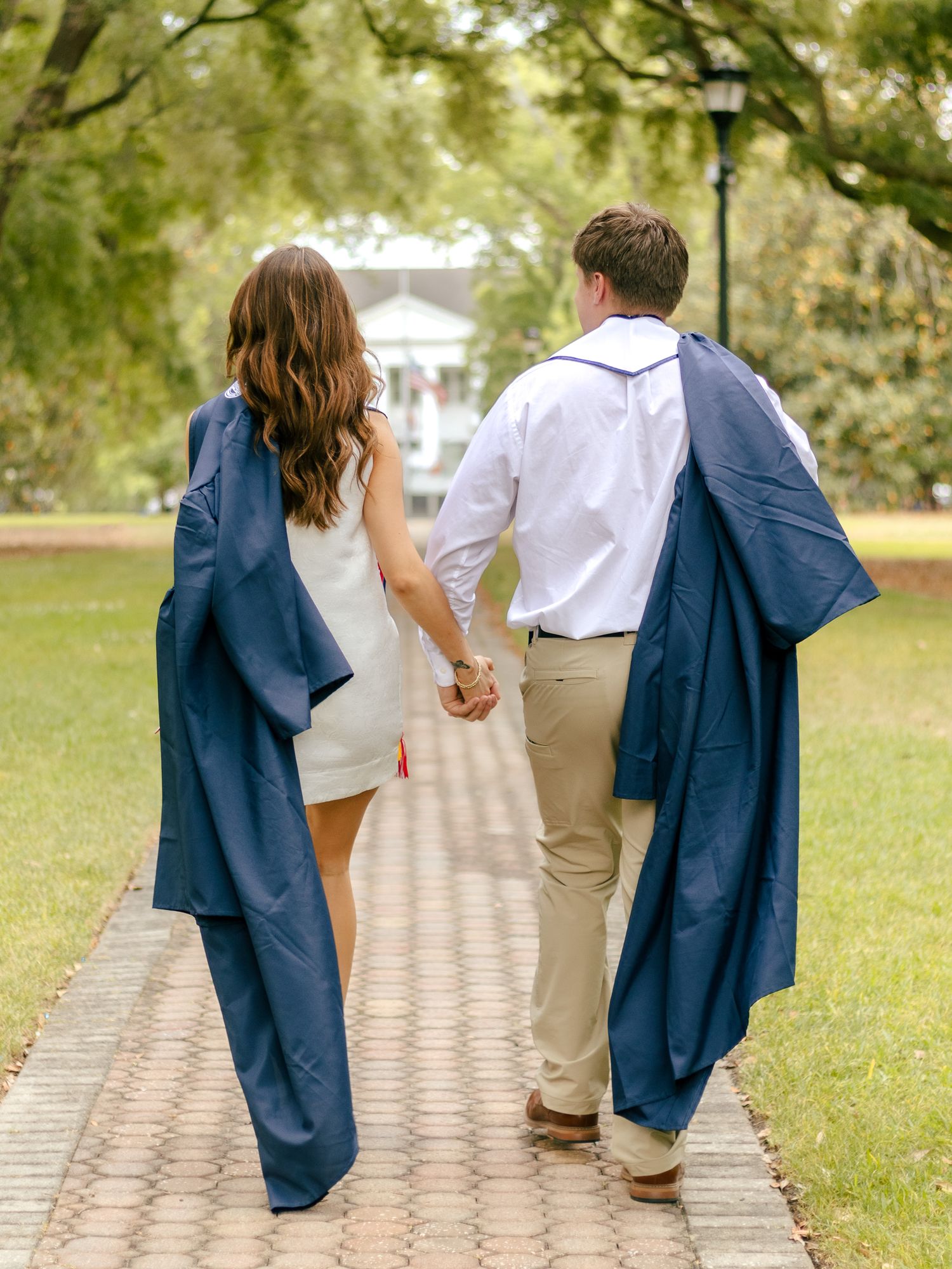 Couple holding hands walking away on a brick path through a sunny park, both carrying navy graduation gowns draped over their shoulders.