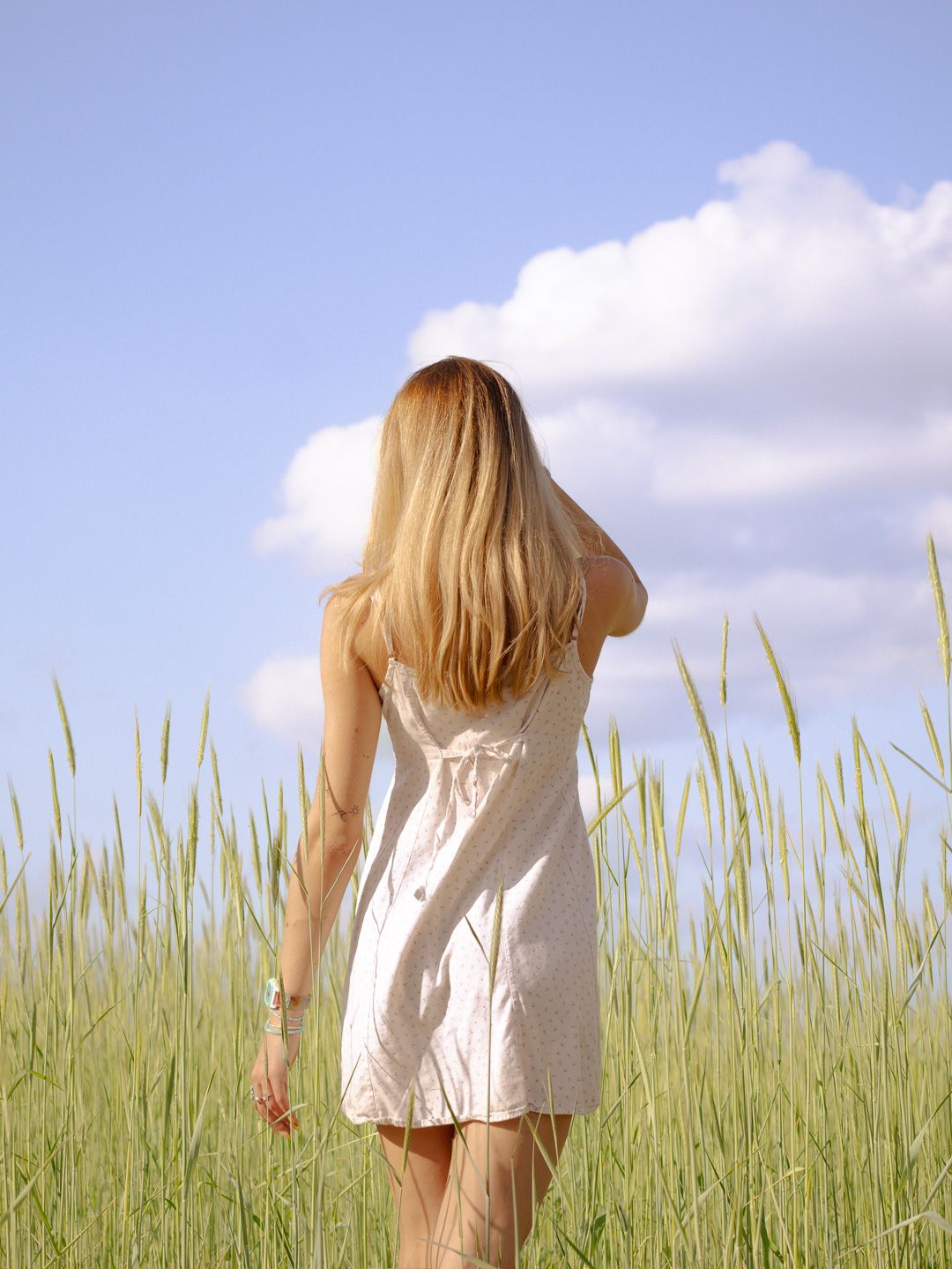 Woman with long blonde hair in a white sundress walks away through a green wheat field under a bright blue sky with fluffy clouds.
