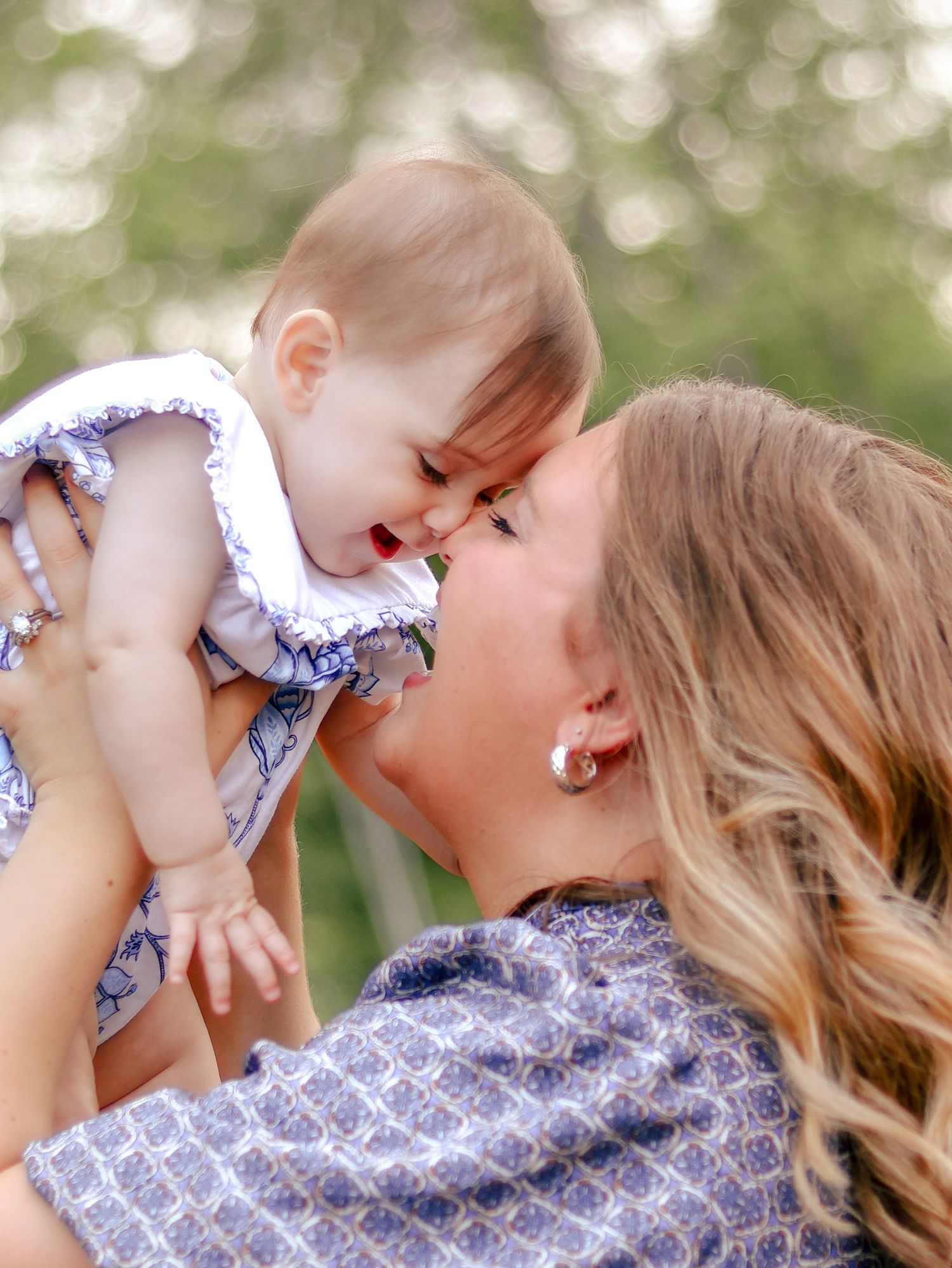 Smiling woman with long blonde hair lifts a happy baby in a white ruffled outfit toward her face outdoors.