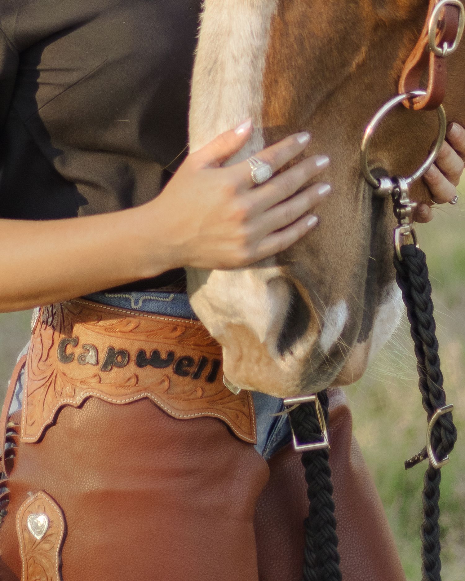 Close-up of a woman's hand with a silver ring resting on a horse's muzzle, beside tooled leather chaps stamped with the name Capwell.