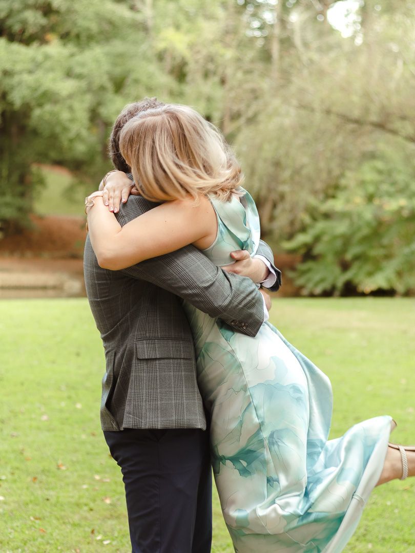 Joyful woman in a teal watercolor dress is lifted off the ground as she hugs her fiancé on a green lawn after his proposal.