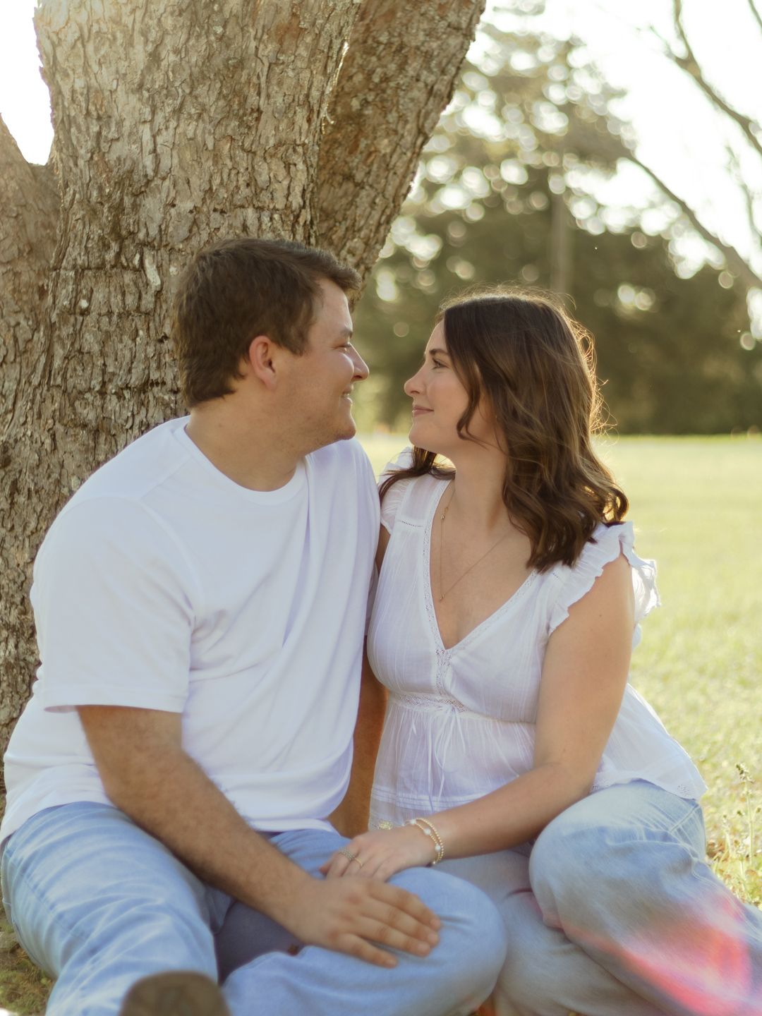A smiling couple in casual white tops and jeans sit close together on the grass beneath a large tree, gazing at each other in warm sunlight.