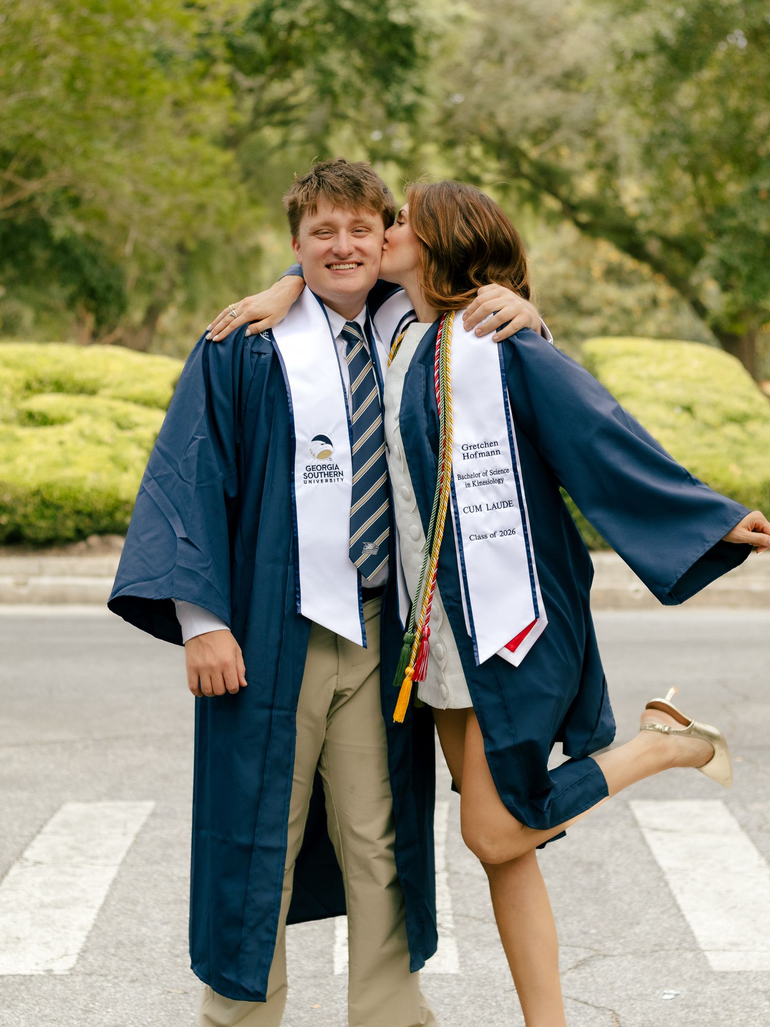 Two graduates in navy caps and gowns with white stoles pose on a crosswalk as one kisses the other's cheek, with green hedges behind.