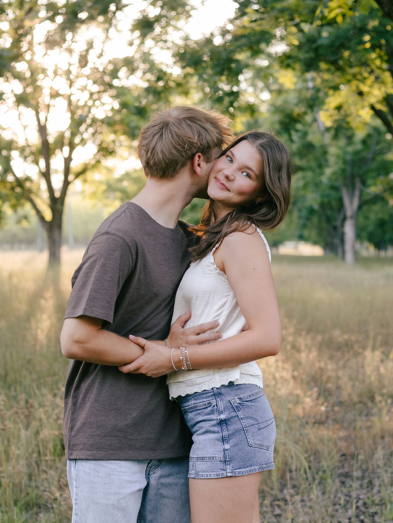 A young couple embraces in a sunlit grassy field with trees, as he kisses her cheek and she smiles warmly.