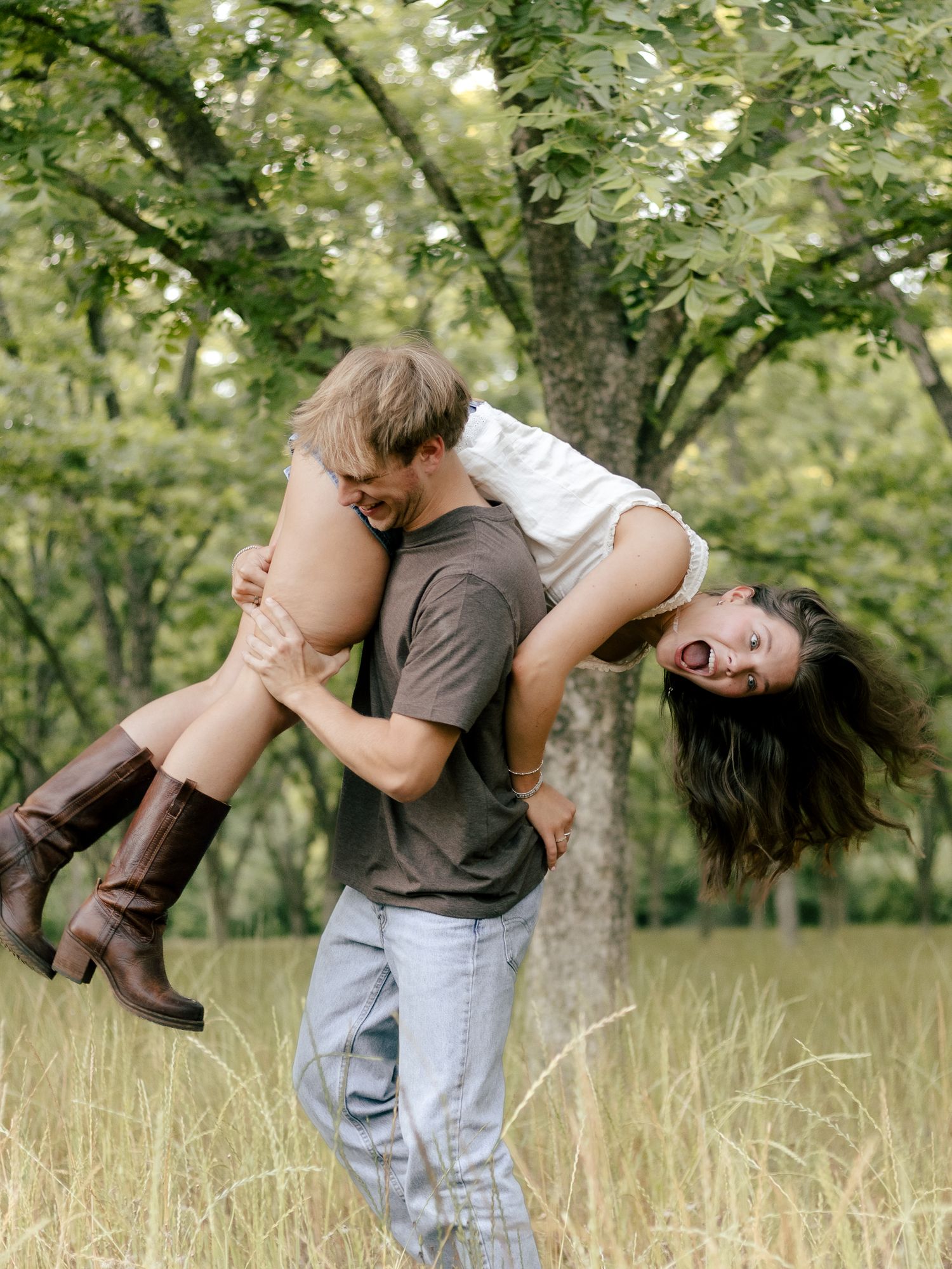 Man in gray t-shirt playfully lifts a laughing woman in brown boots over his shoulder in a sunny grassy field under green trees.