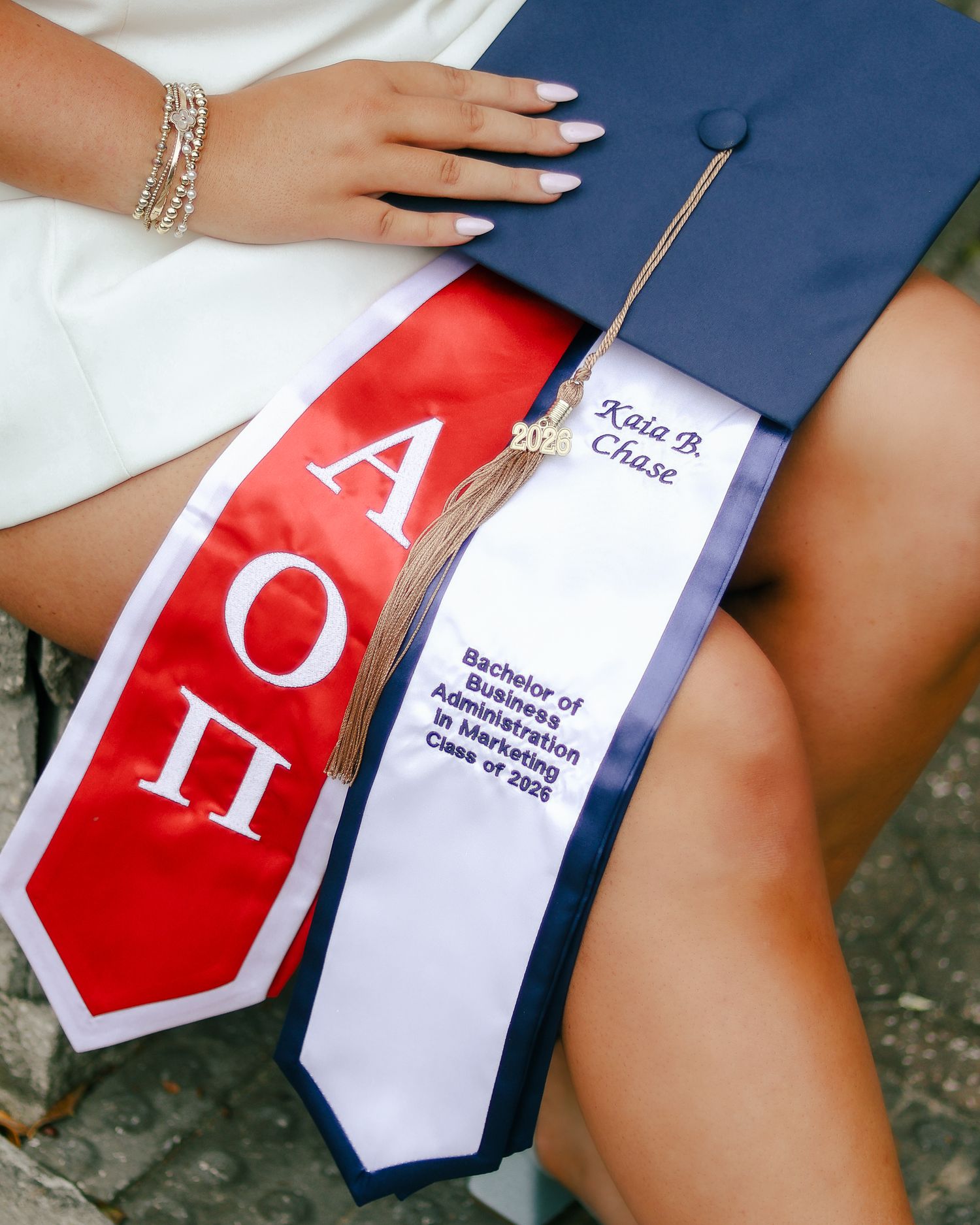 Graduate sitting with a navy cap and gold tassel, wearing a red Alpha Omicron Pi sash and a white marketing business degree stole.