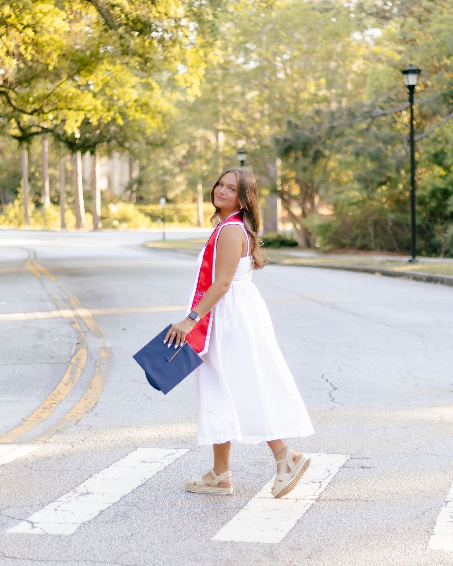 Woman in a white sundress and red sash walks across a crosswalk on a tree-lined street, holding a navy blue tote bag.