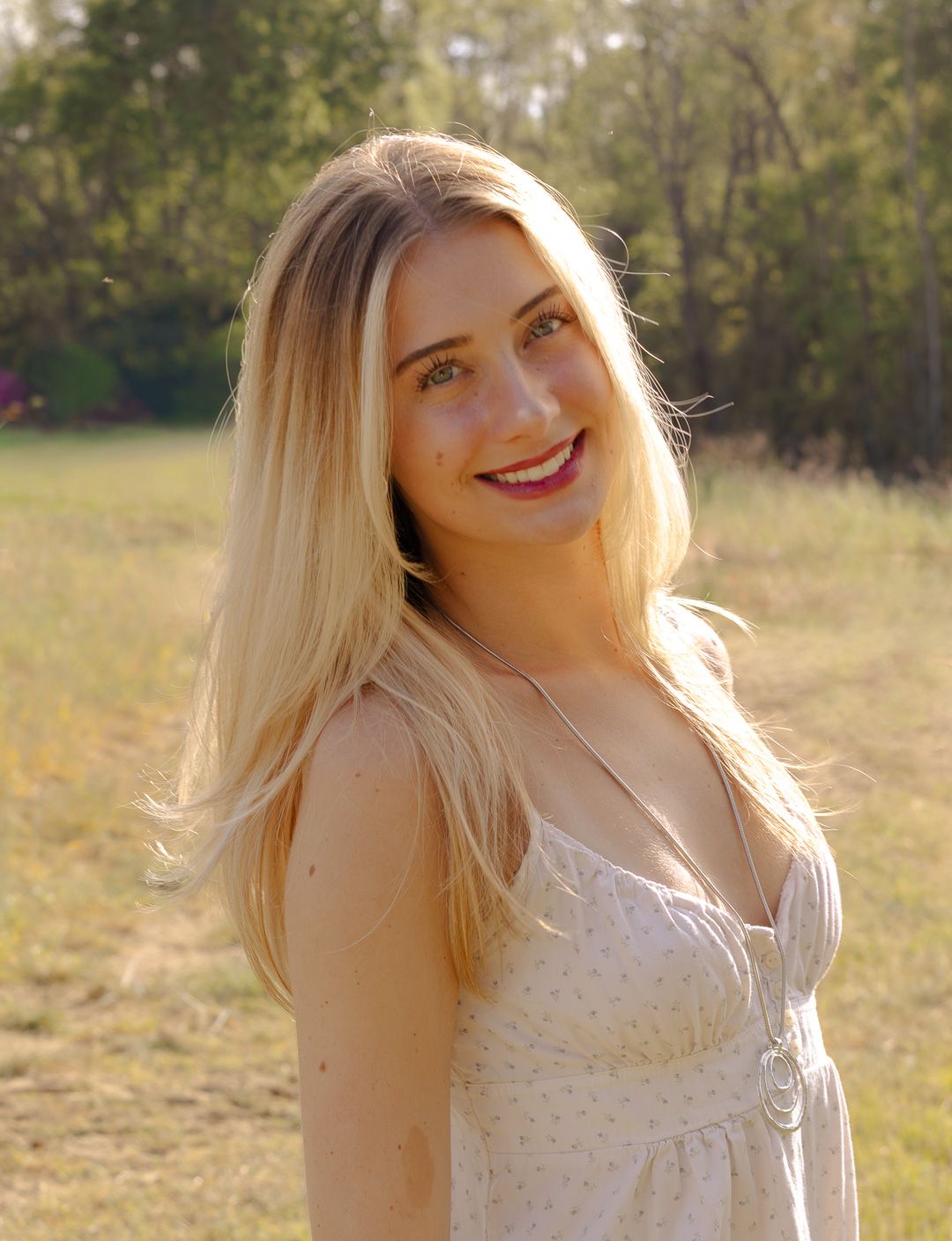 Smiling woman with long blonde hair in a white sundress and pendant necklace standing in a sunlit grassy field with trees behind.