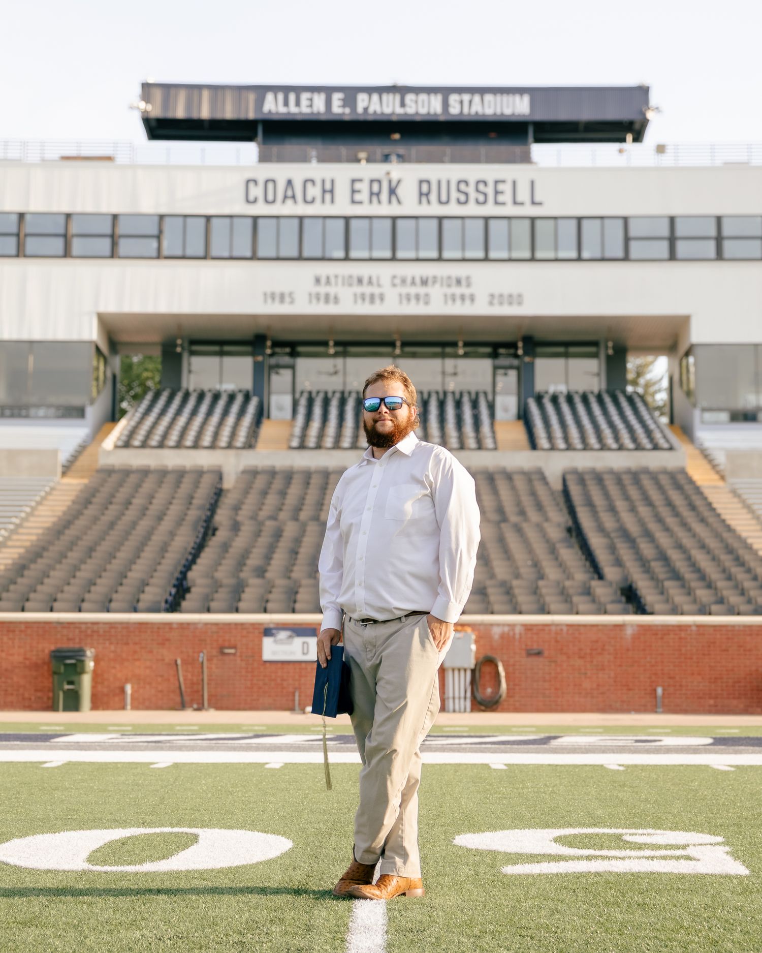 Man in sunglasses, white dress shirt and khaki pants stands on the field at Allen E. Paulson Stadium, with empty stands behind him.
