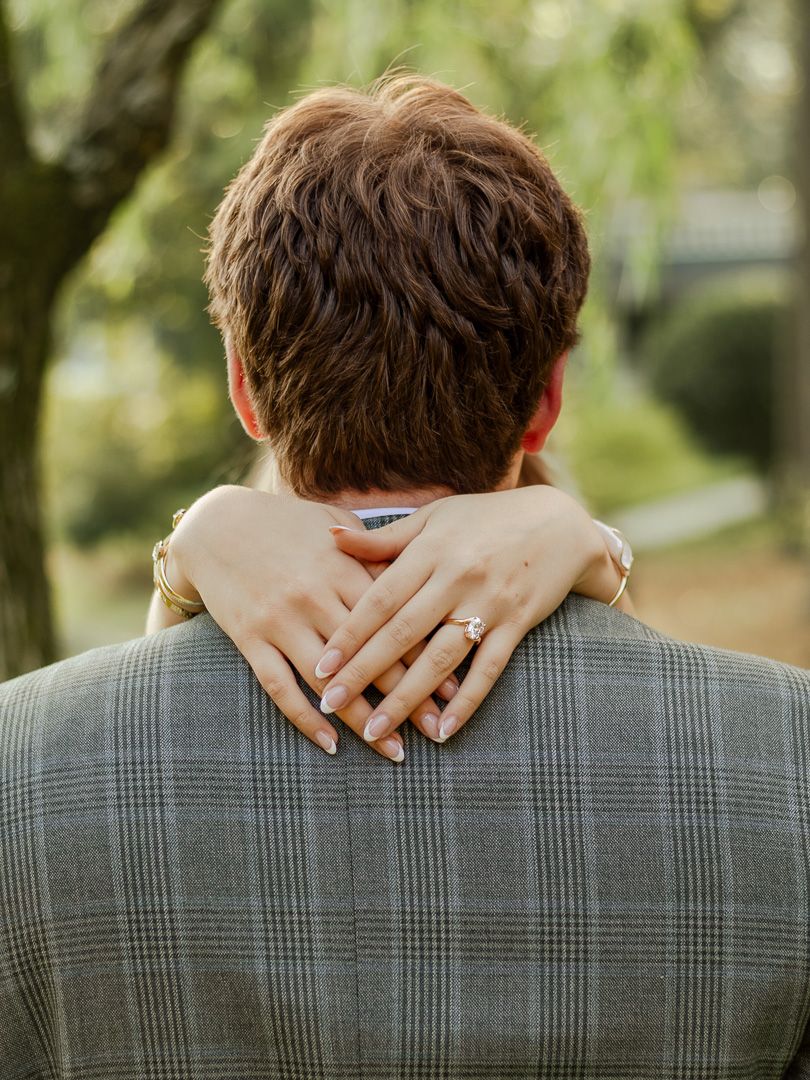 A woman wraps her arms around a man's shoulders, her sparkling engagement ring visible against his gray plaid jacket.