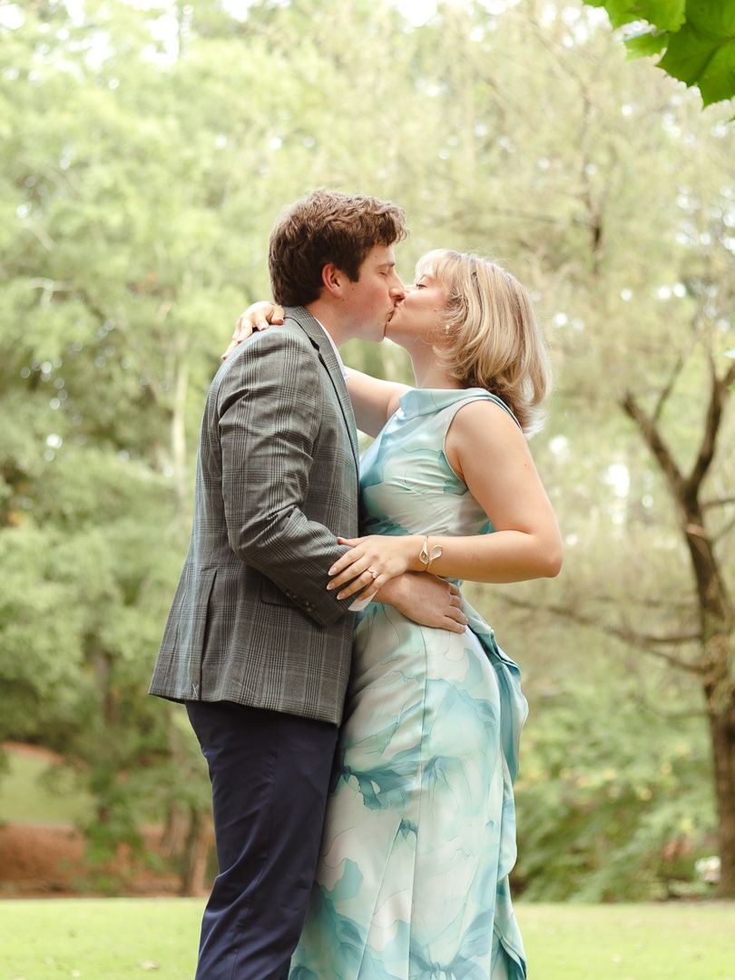 A couple shares a kiss in a sunny park, he in a gray plaid blazer and she in a flowing blue watercolor dress, surrounded by green trees.