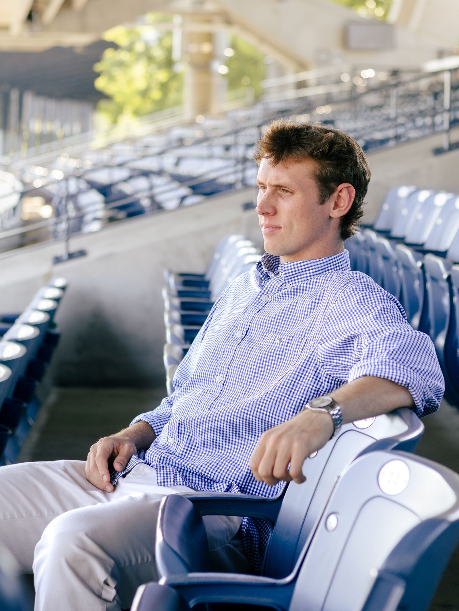 Young man in a blue gingham button-up shirt and khaki pants sits in a blue stadium seat, gazing into the distance.