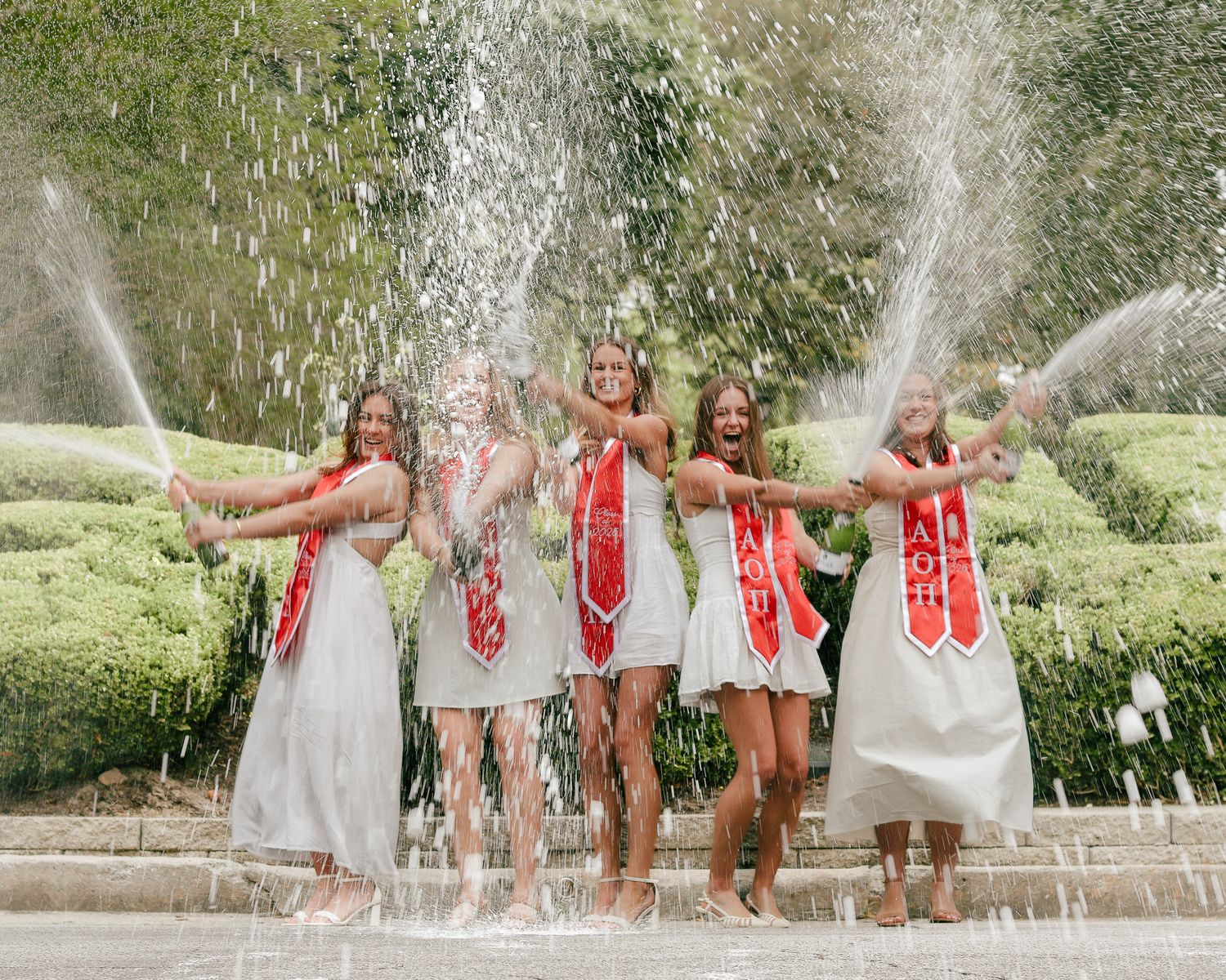 Five women in white dresses and red graduation sashes laugh and pose barefoot as fountain water sprays around them in a garden.
