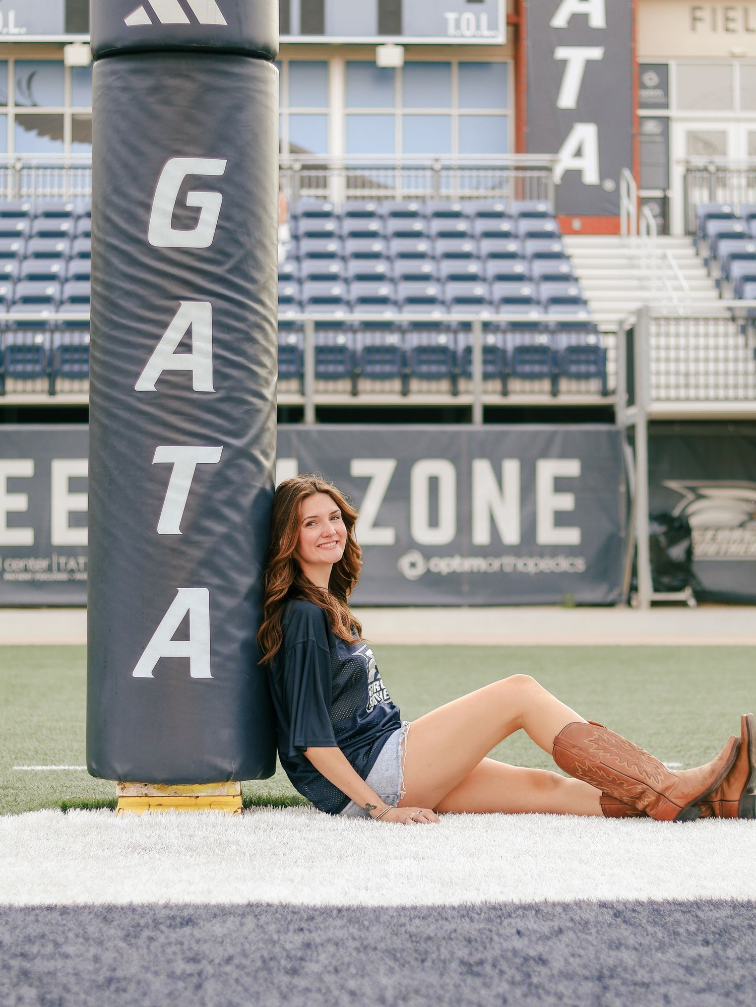 Young woman in cowboy boots and a dark t-shirt sits on turf leaning against a padded goalpost at a football stadium with empty bleachers.