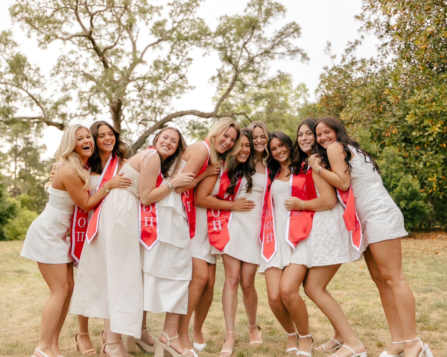 Group of smiling women in white dresses and red sashes pose arm in arm outdoors under trees in a sunny park, celebrating graduation.