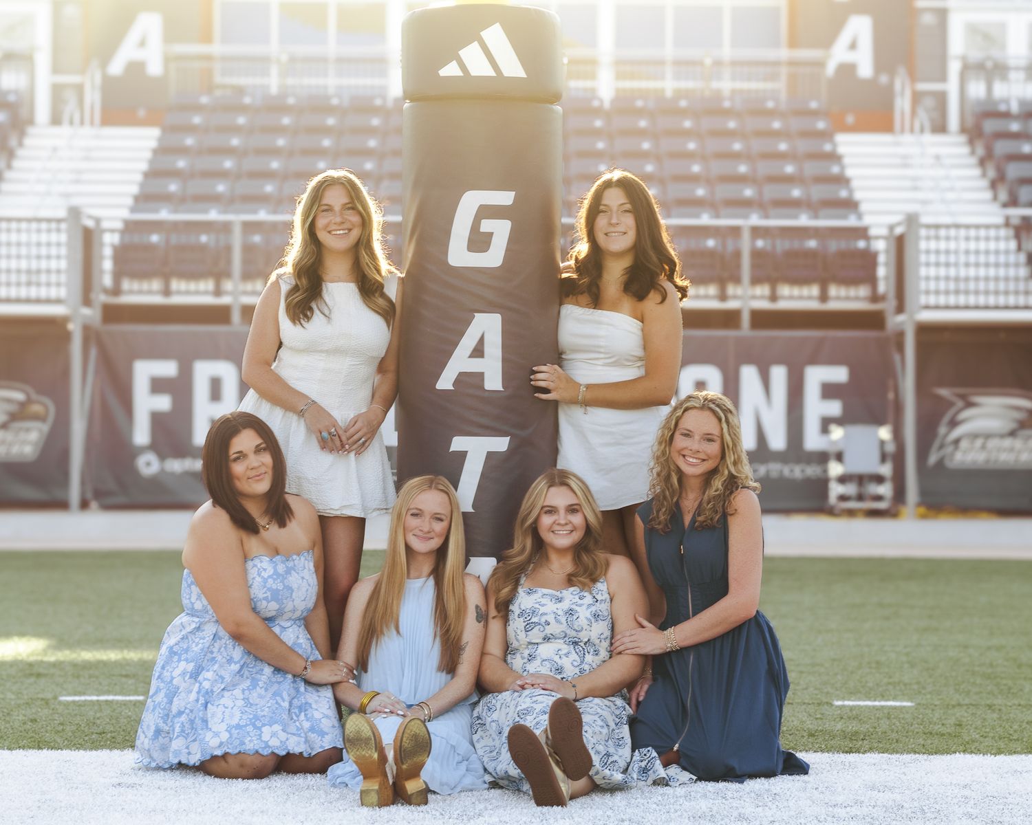 Five young women in white and blue dresses pose together on a football field in front of a stadium goalpost pad and bleachers.