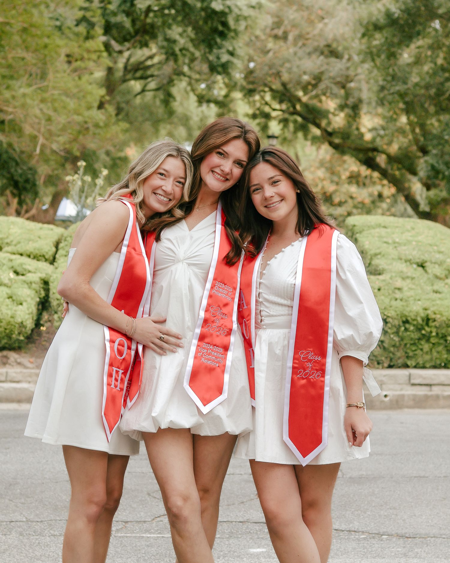 Three smiling women in white dresses and red graduation sashes pose arm in arm outdoors on a tree-lined street.