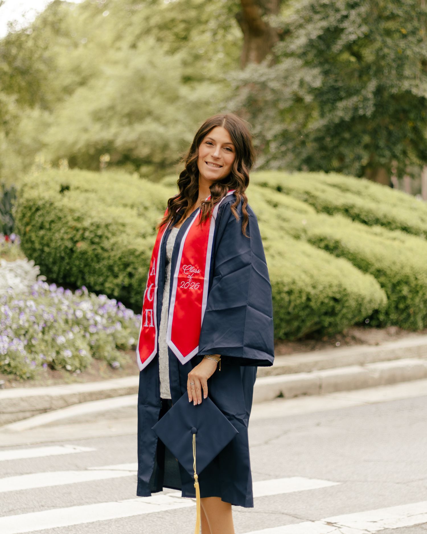 Smiling graduate in a navy cap and gown with a red stole holds her mortarboard while posing on a crosswalk near green hedges.