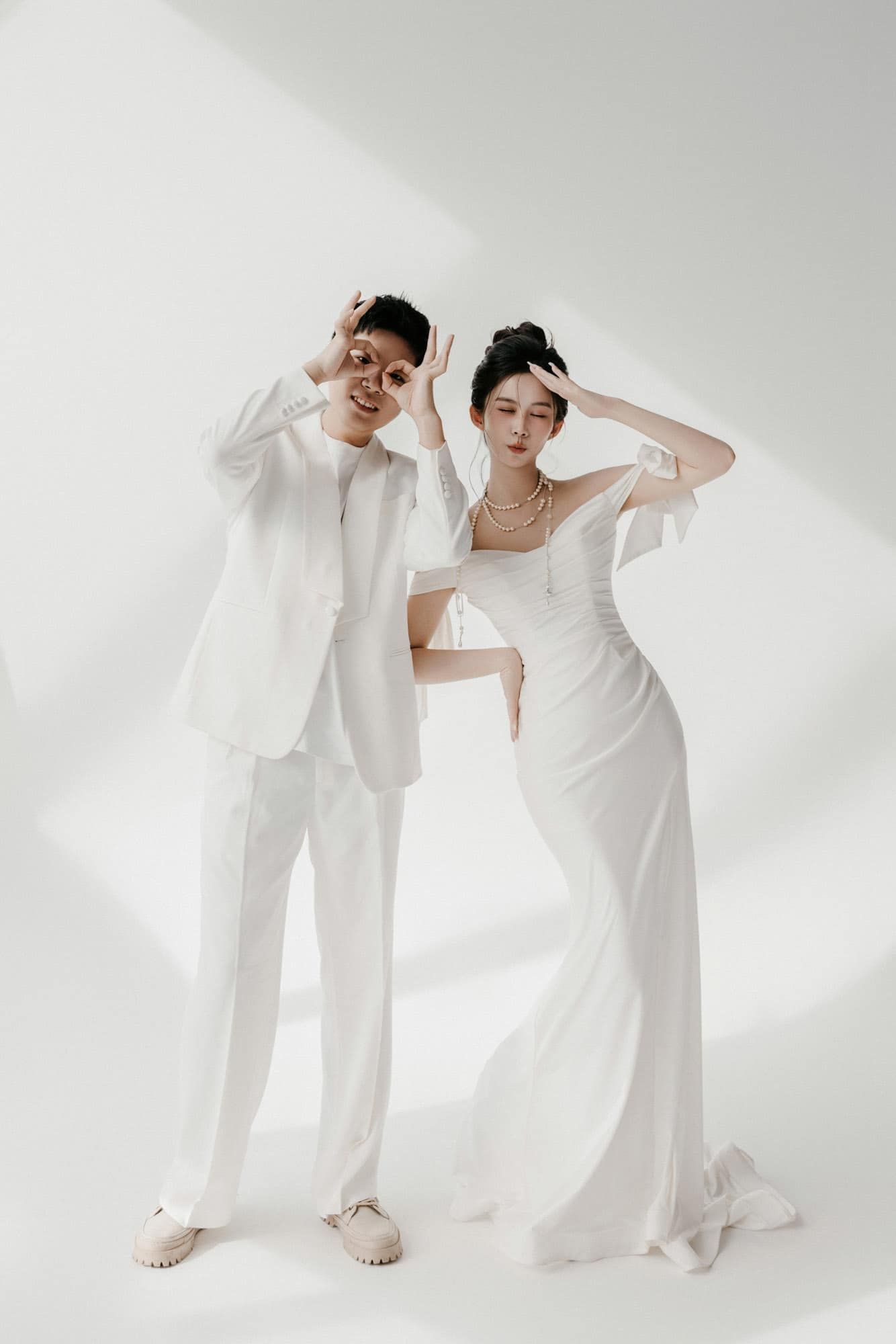 A couple in matching white outfits pose playfully in a studio, the bride in a flowing gown and groom in a suit.