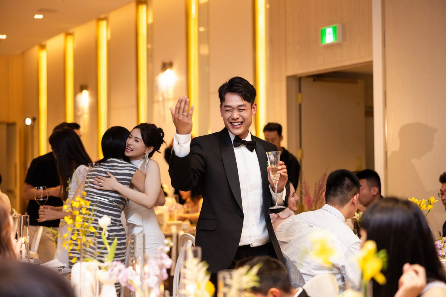 Cheerful groom in tuxedo waving and holding champagne glass at lively wedding reception.