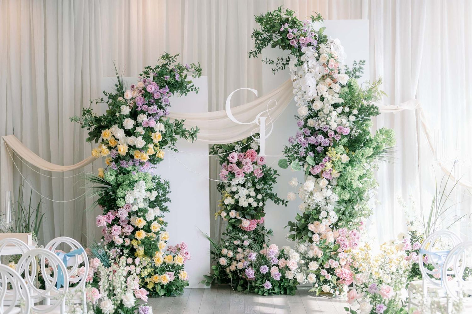 Bright wedding ceremony backdrop with two asymmetric floral arches covered in pastel blooms and greenery.