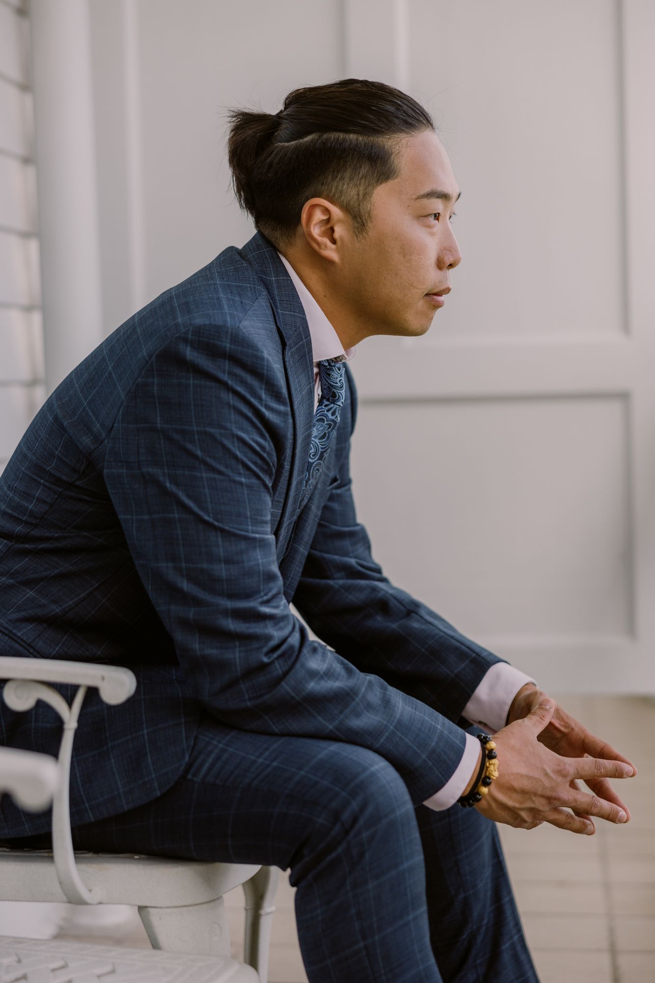 Asian man in navy plaid suit sitting thoughtfully on white chair in elegant setting.