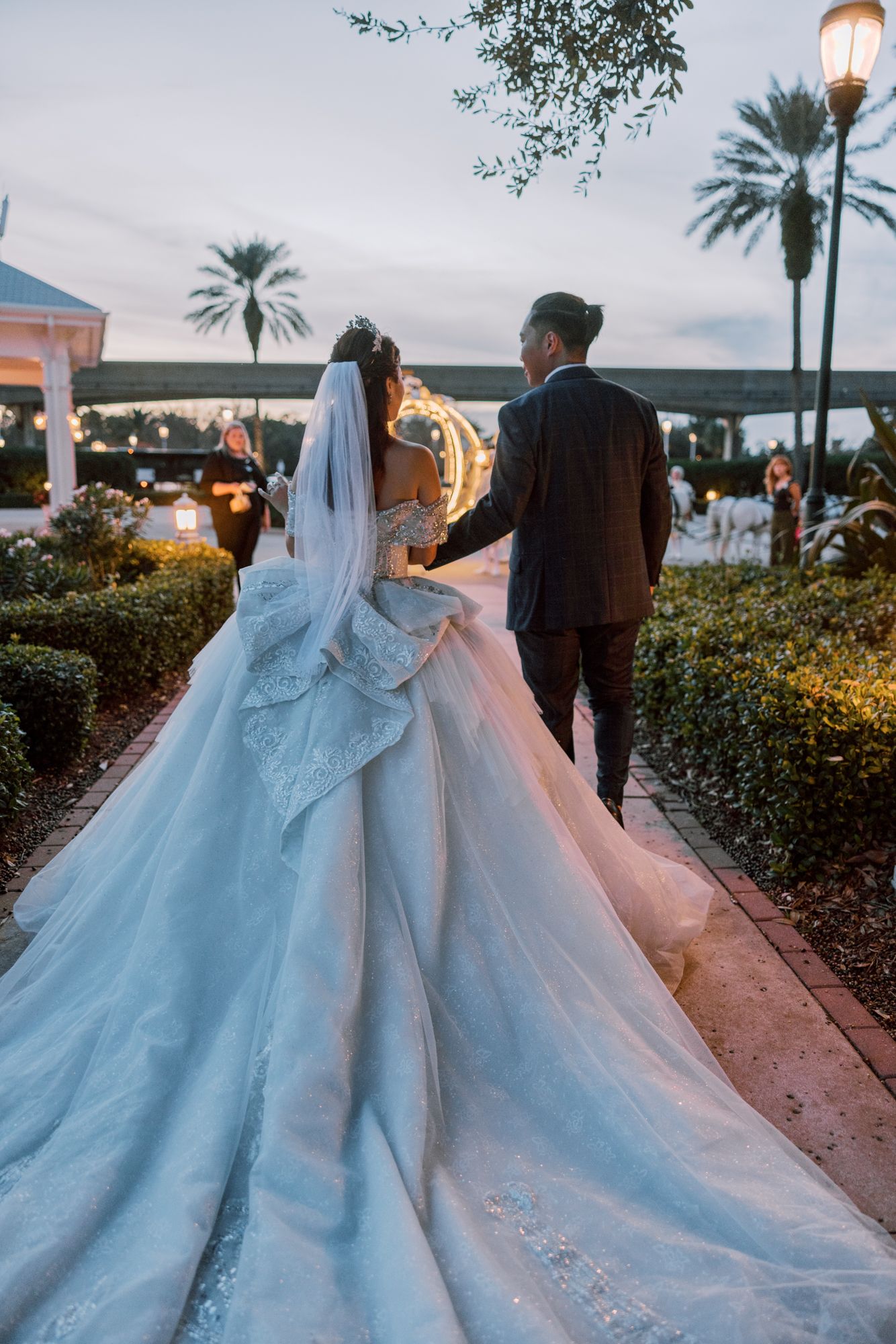 Bride in stunning white ball gown with long train walks hand-in-hand with groom at twilight garden venue.