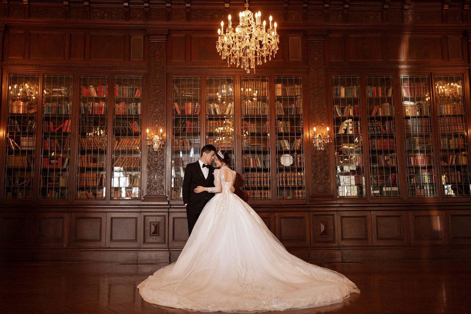 Bride and groom embracing in front of floor-to-ceiling bookshelves under a chandelier in a grand library.