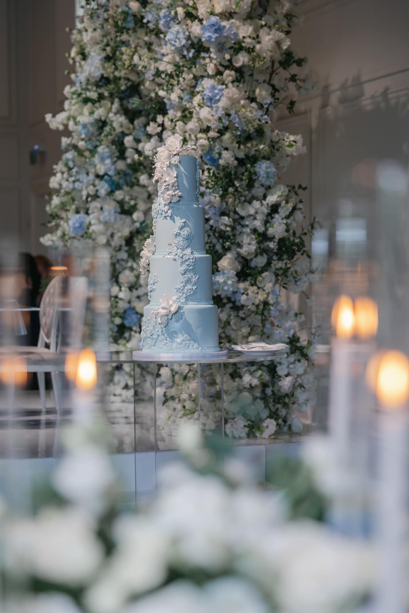 Three-tier light blue wedding cake with white floral appliques displayed in front of a blue and white floral column.