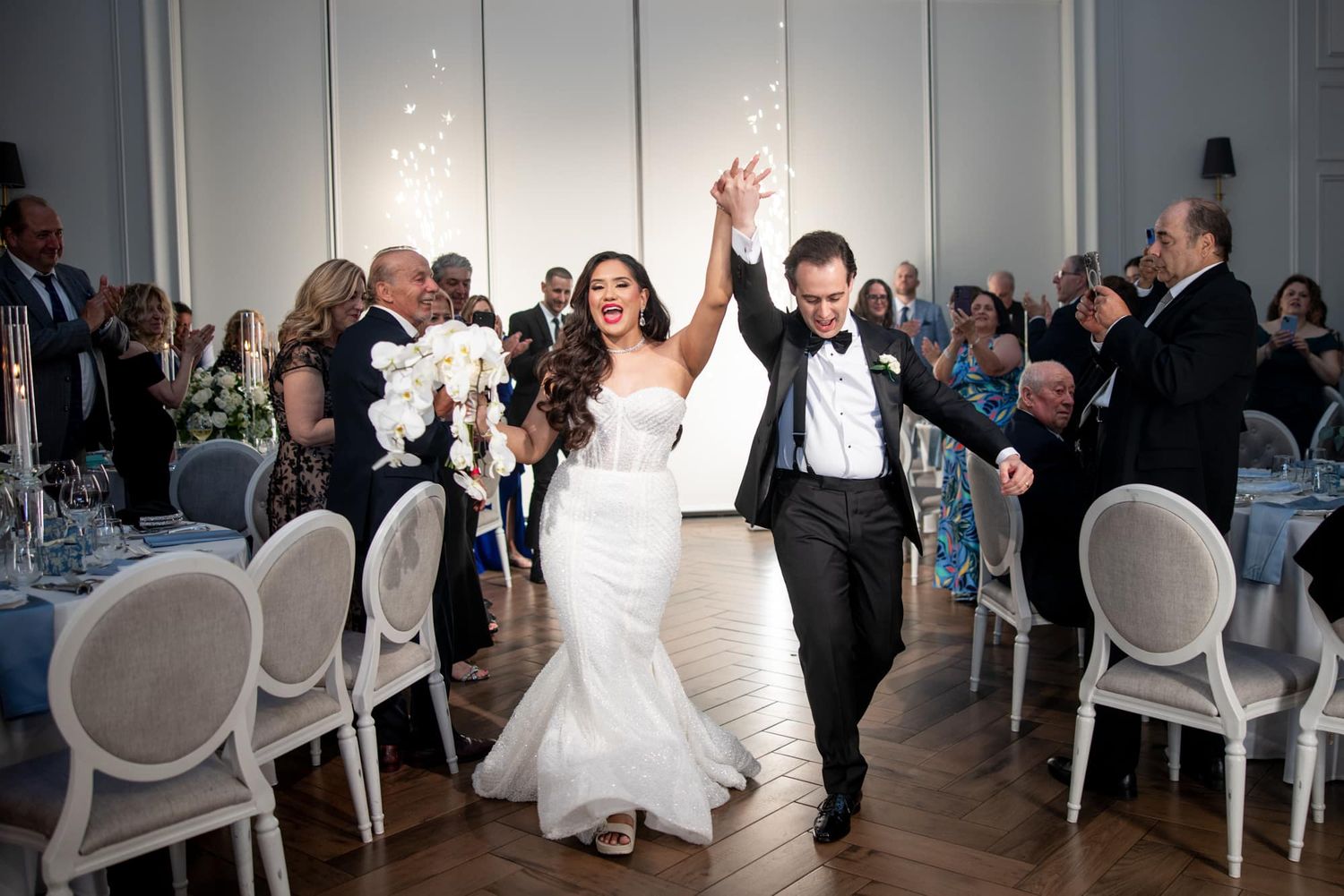 Joyful bride and groom raising hands during grand entrance at elegant wedding reception with guests applauding.