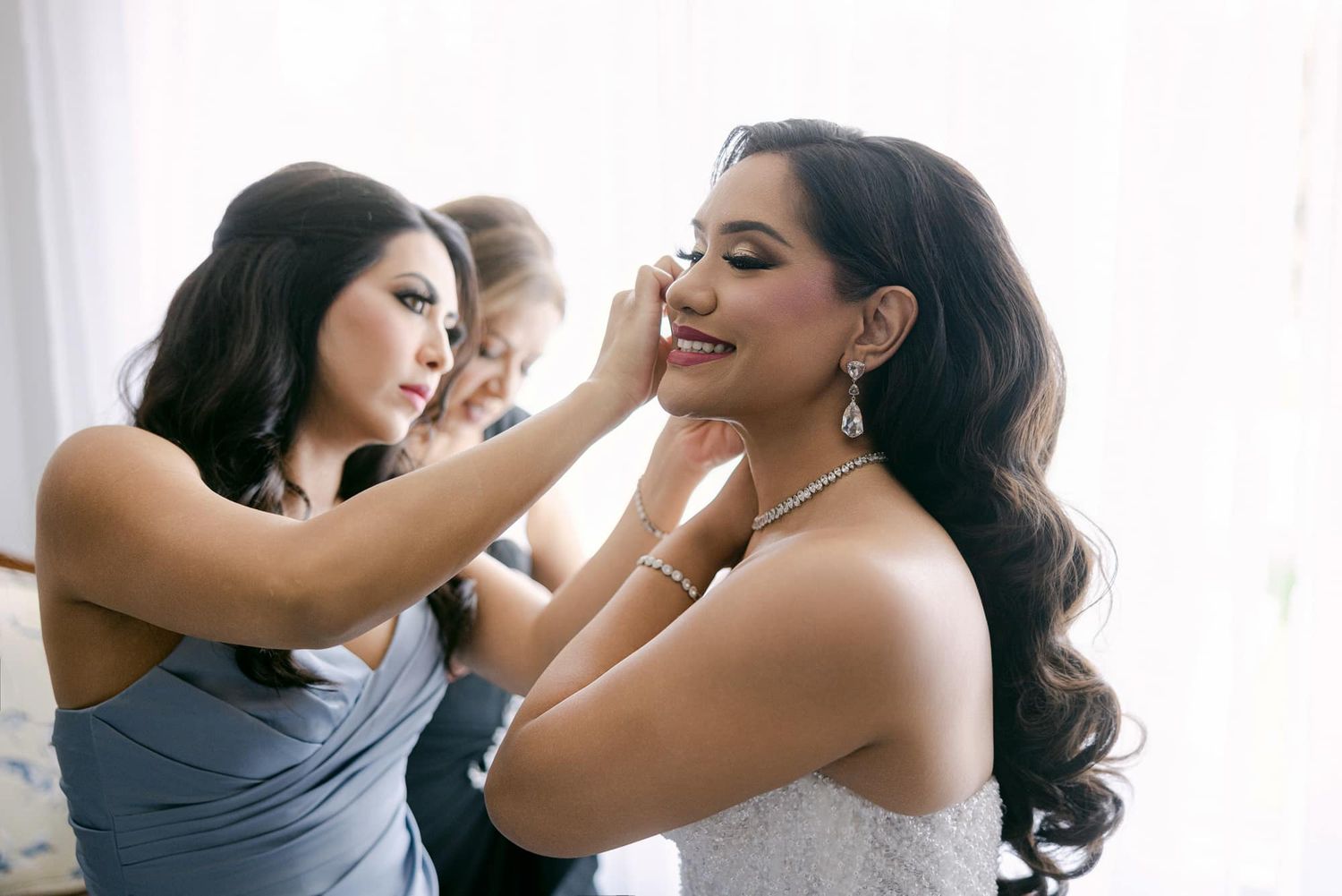 Bridesmaids helping a smiling bride with her makeup and jewelry while getting ready for her wedding.