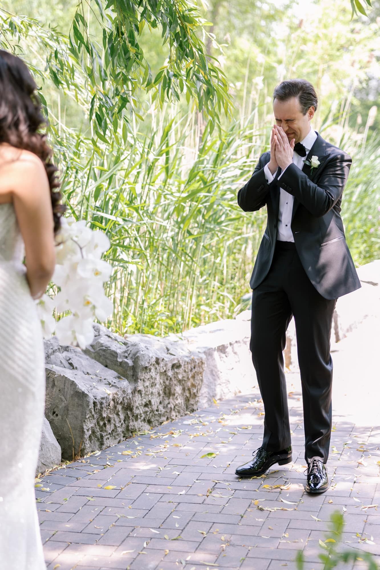 Groom in black tuxedo clasping hands in awe during first look at outdoor wedding ceremony.