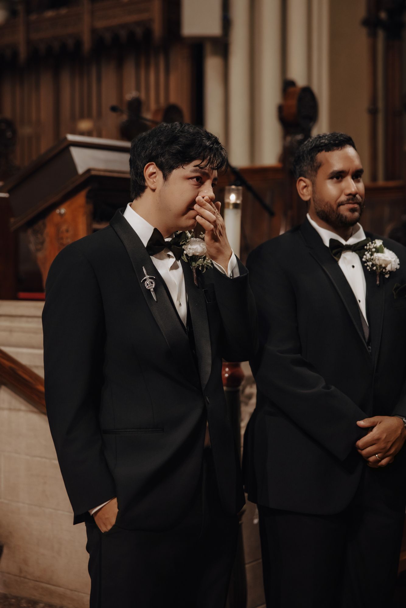 Emotional groom in a black tuxedo covers his mouth as he waits at the church altar beside a smiling groomsman wearing a boutonniere.