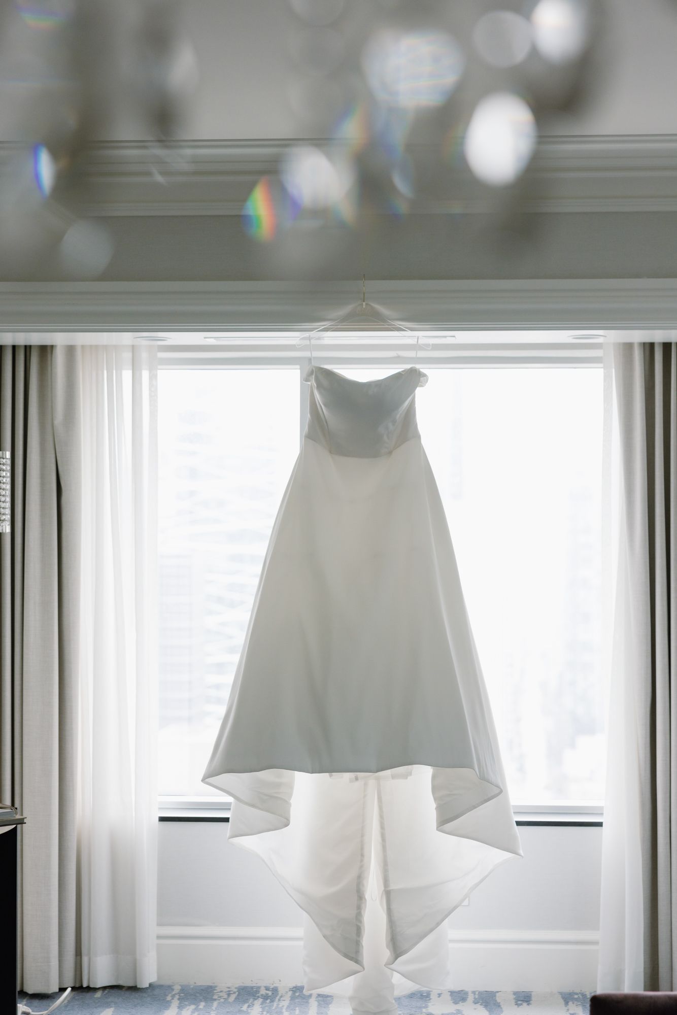 A strapless white wedding gown with a long train hangs in front of a bright window with sheer curtains beneath a crystal chandelier.