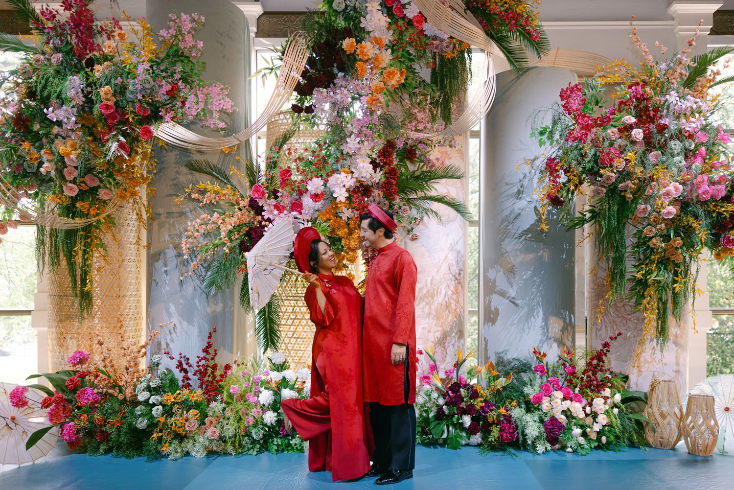 Couple in matching red traditional outfits stand together beneath arches of colorful floral arrangements at an elegant wedding ceremony.
