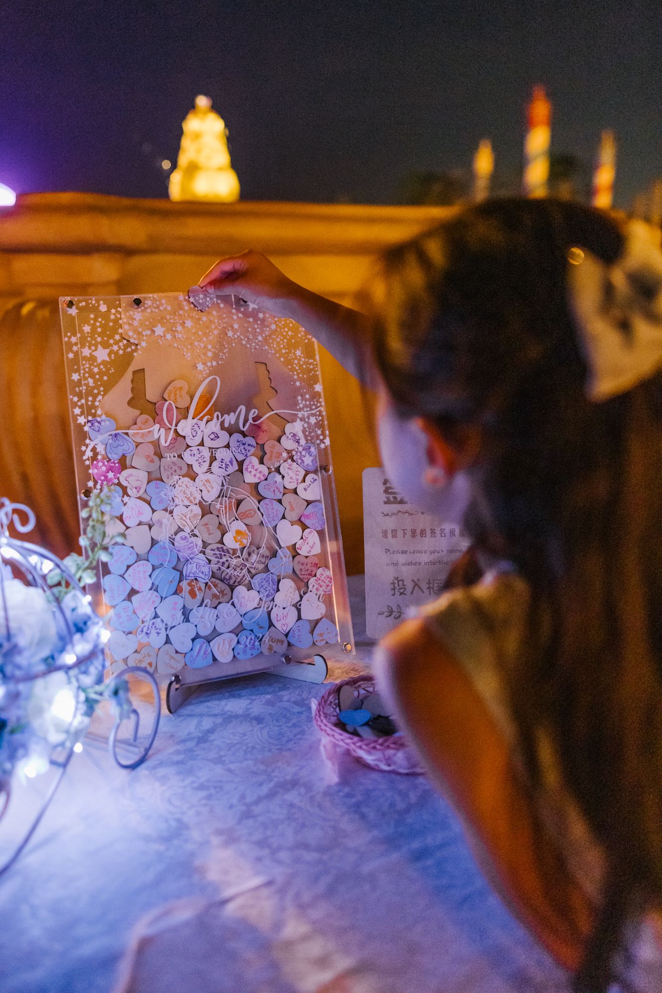 Woman holding a floral drop-top wedding guest book sign at an outdoor nighttime celebration.