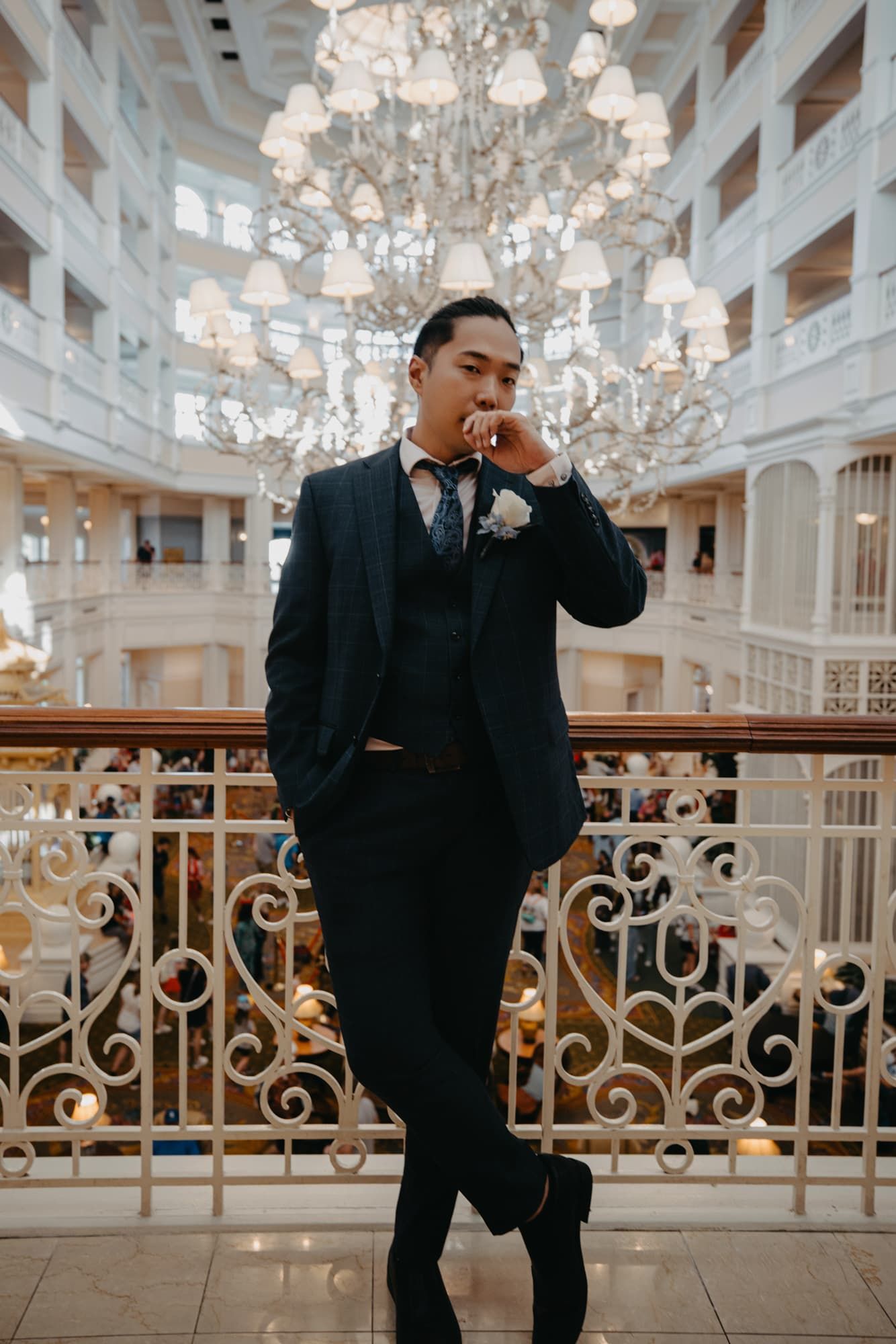 Dapper groom in navy suit with boutonniere poses confidently on balcony of elegant chandelier-lit grand hall.
