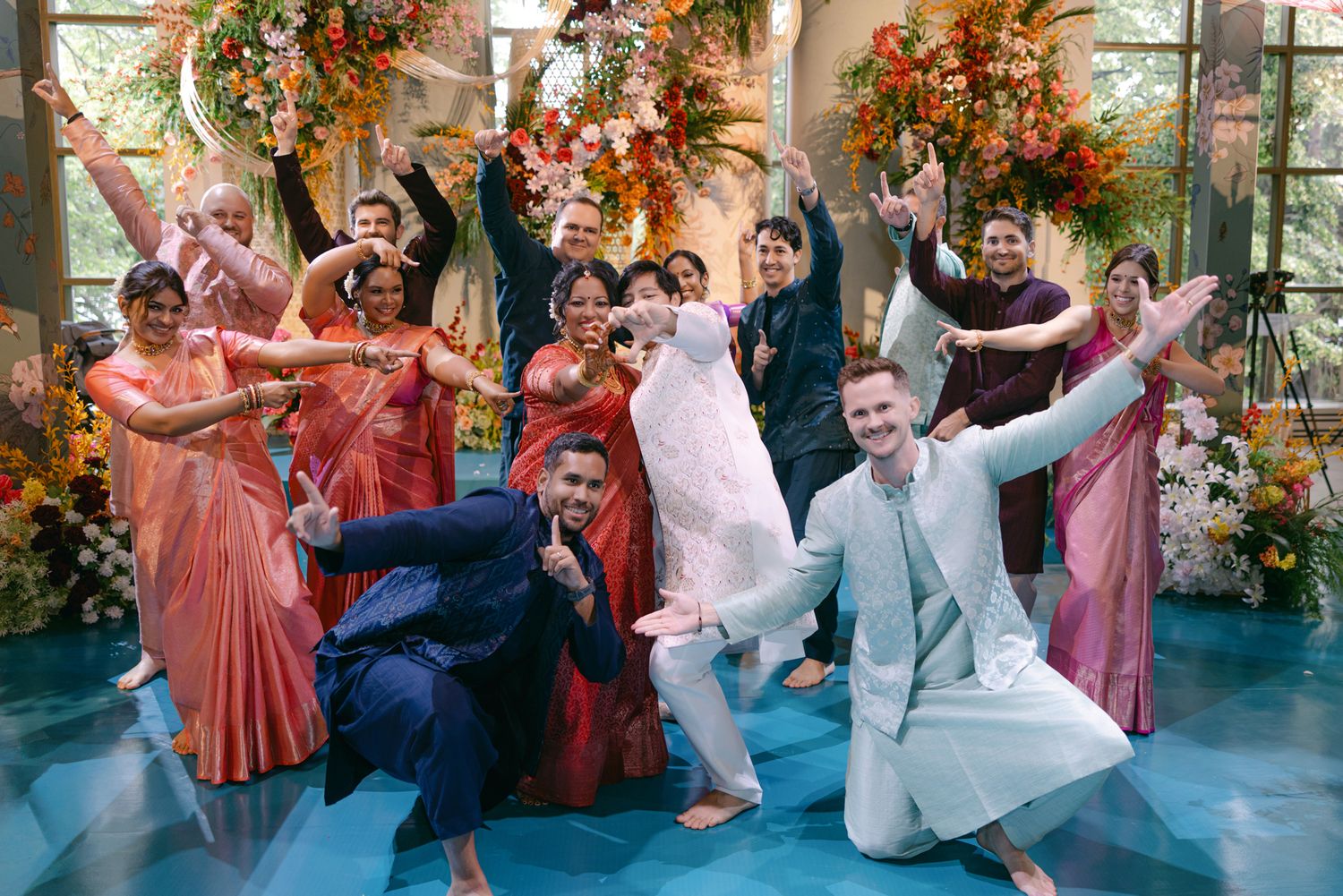 Group of smiling people in colorful traditional Indian attire and formal suits dancing joyfully together in a floral-decorated hall.