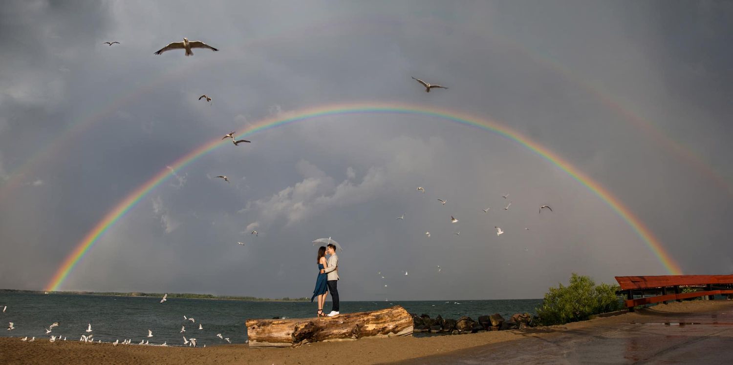 Couple embracing under a stunning full rainbow over a lakeside landscape.