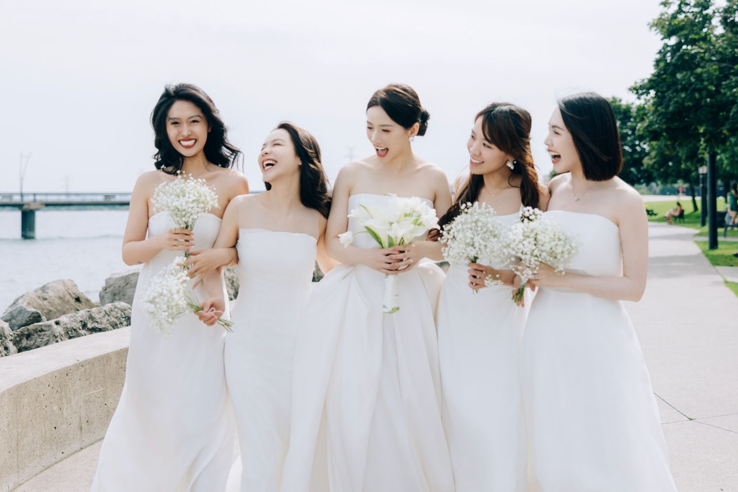 Five bridesmaids in white dresses laugh joyfully outdoors holding floral bouquets.
